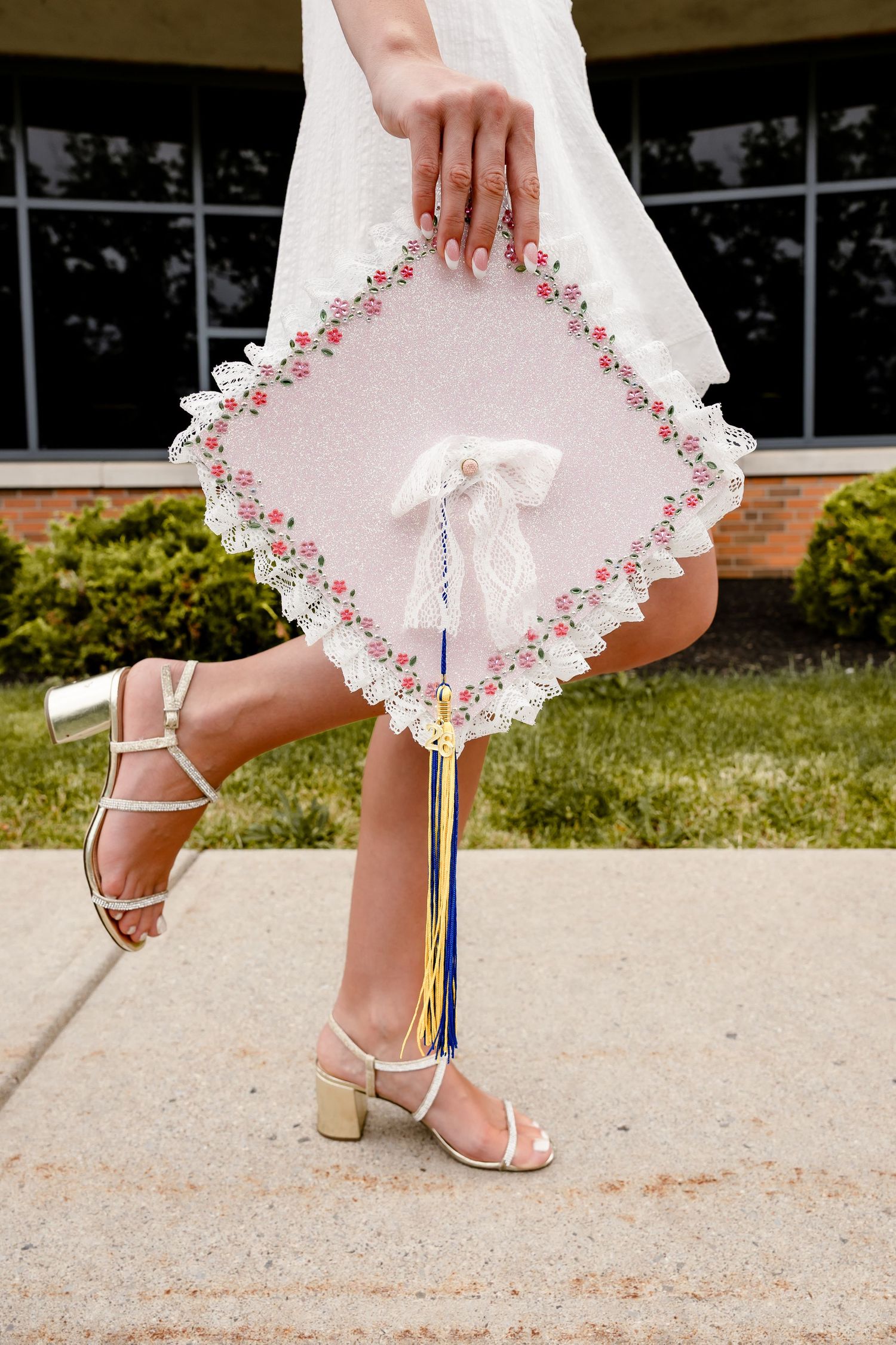 Graduate holding decorated floral mortarboard cap with colorful tassels, wearing white dress and gold heeled sandals outdoors.