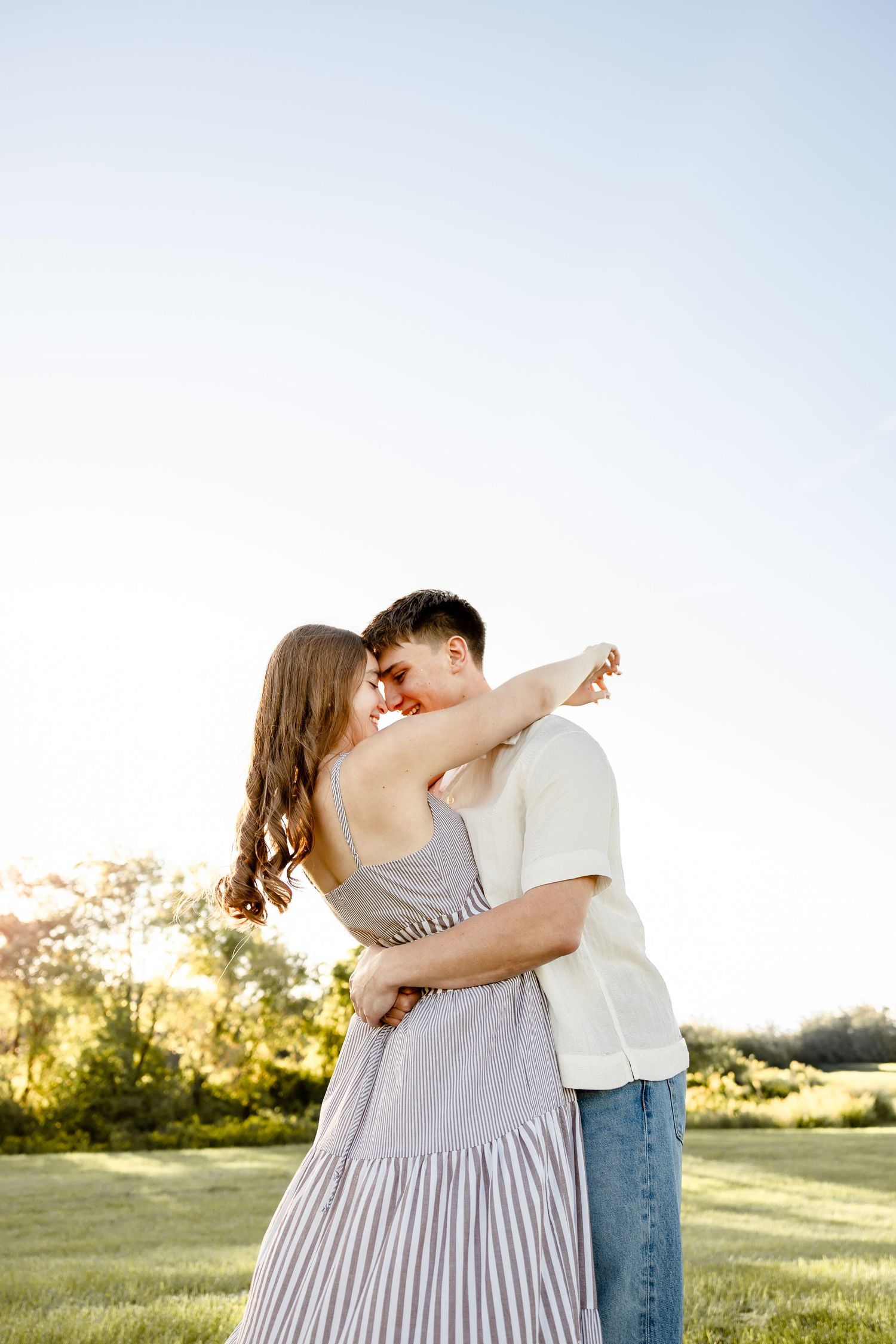 A young couple embraces tenderly in a sunny green field, the woman in a striped dress and man in white shirt and jeans.