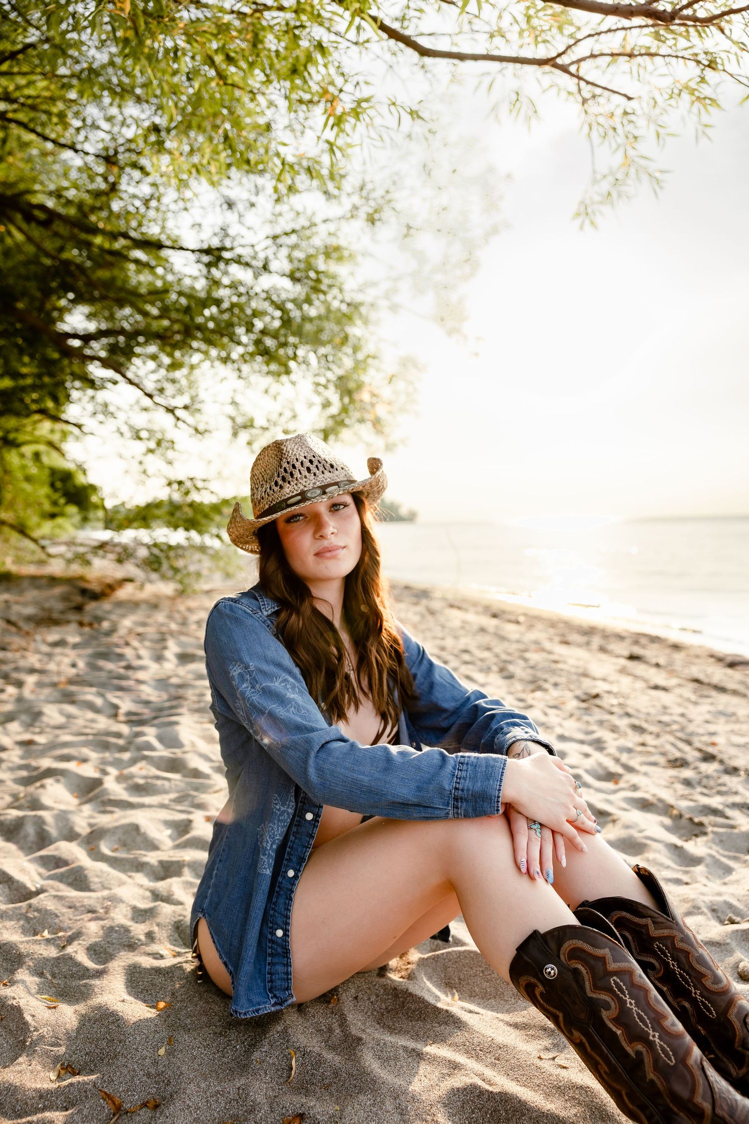 Young woman in denim shirt, cowboy hat, and boots sitting on sandy beach under trees.
