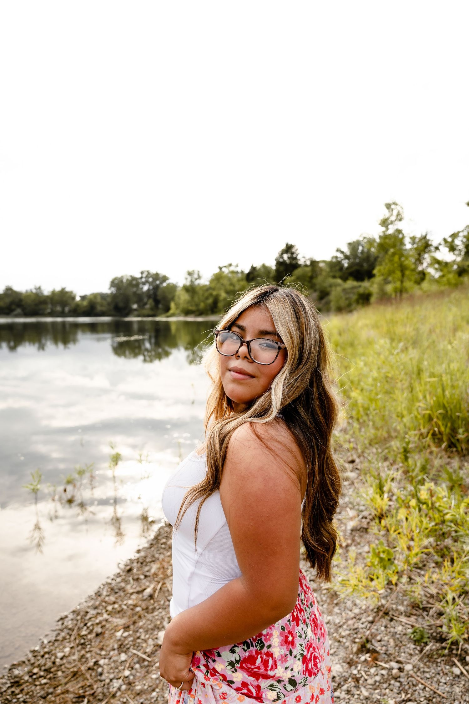 Young woman with long hair and round sunglasses posing by a serene lake shore wearing a white top and floral skirt.