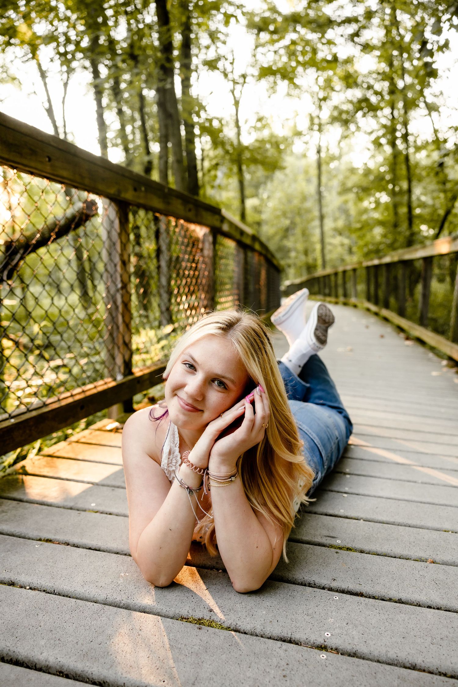 Smiling blonde teen girl lying on a wooden bridge, talking on phone, surrounded by lush green forest trees.