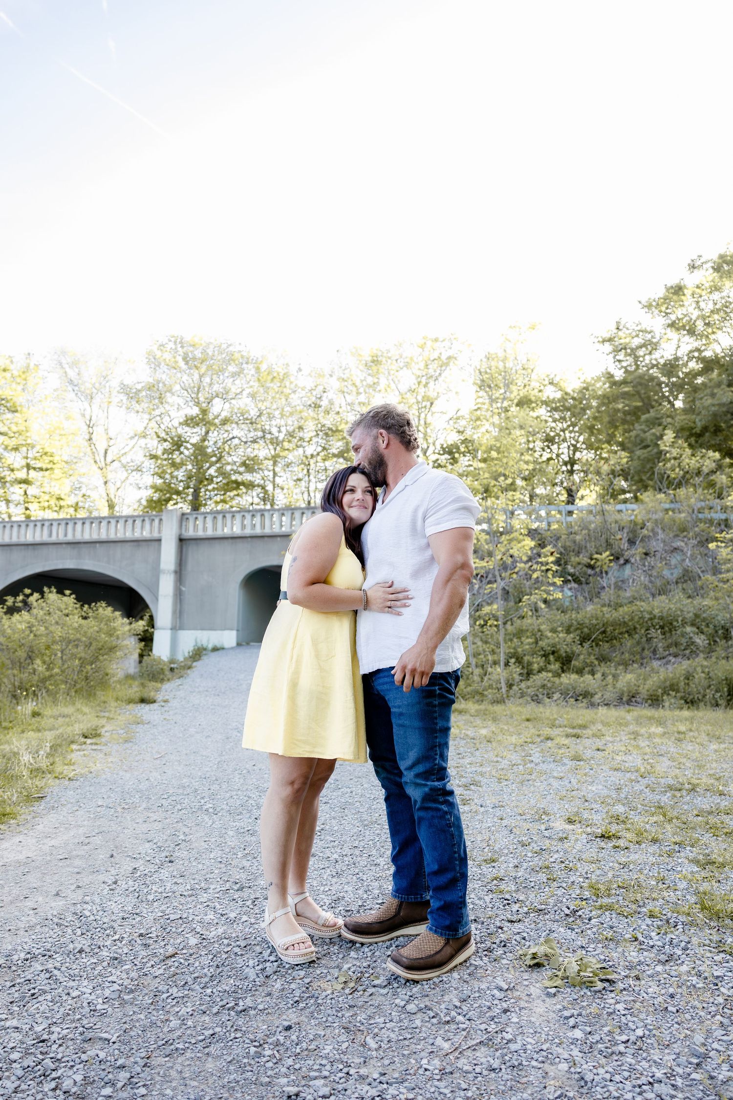 Couple embracing outdoors near a stone bridge, woman in yellow dress and man in white shirt and jeans.