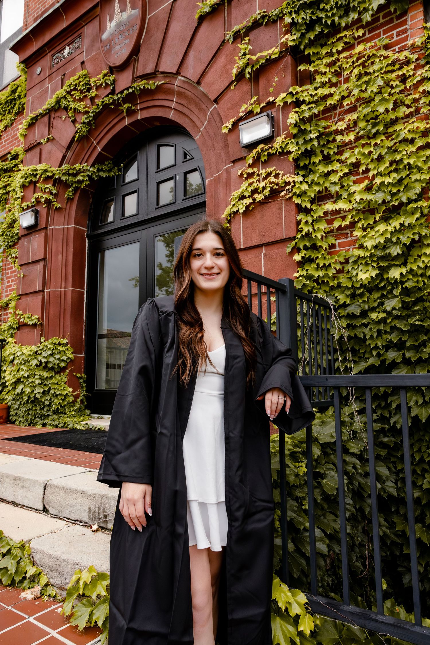 A smiling young woman in a black graduation gown and white dress poses outside an ivy-covered red brick building.