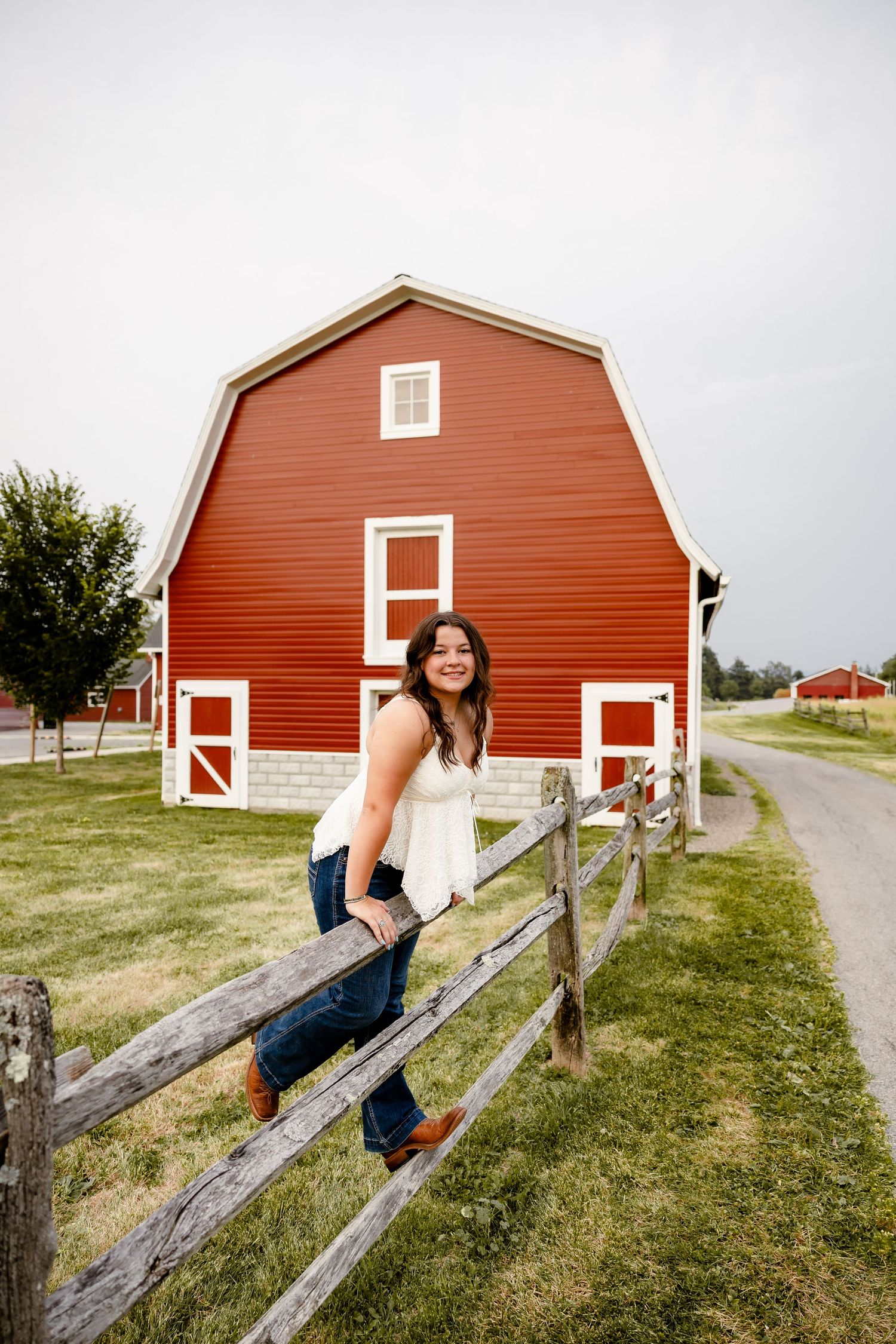 Young woman in white top and jeans leaning on wooden fence with red barn in background on sunny farm.