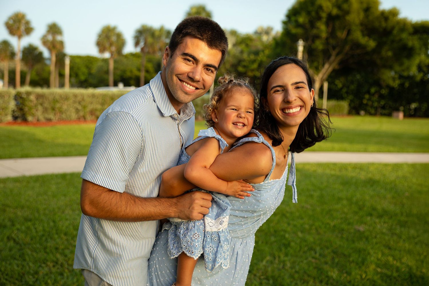 Happy family of three posing in a sunny park, parents holding their smiling toddler daughter.