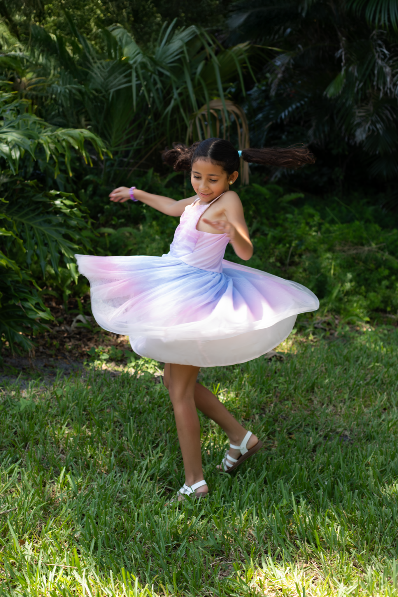 A young girl in a pastel rainbow dress twirls outdoors in a lush green garden.