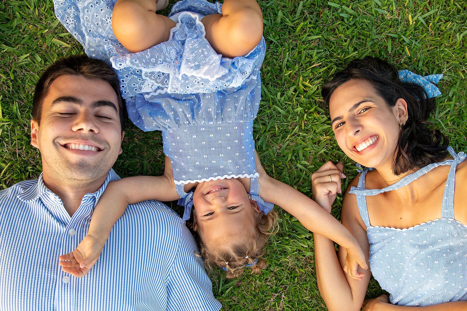 Smiling family of three lying on green grass looking up at camera in blue outfits.