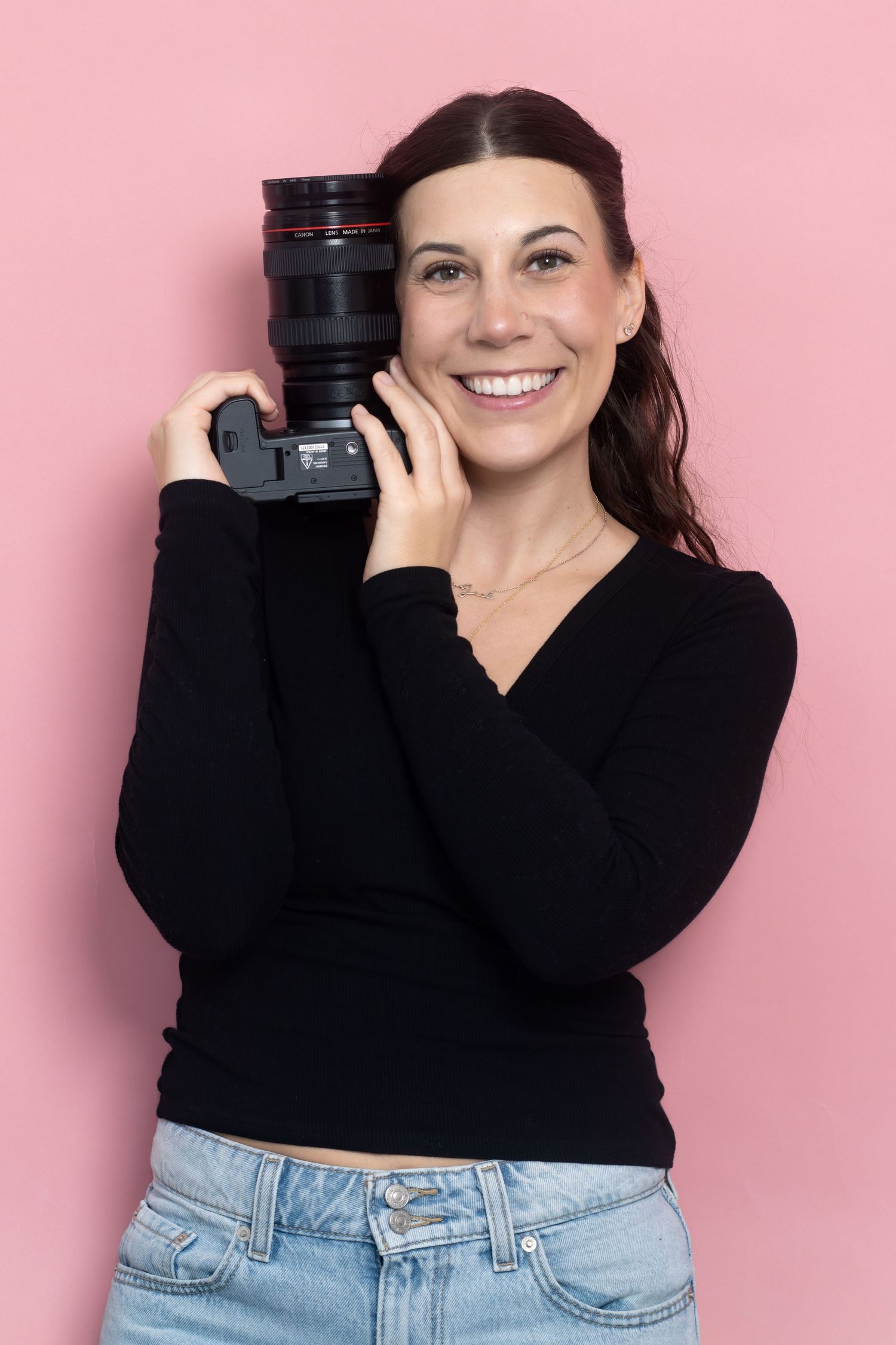 Photographer in black sweater and jeans poses with camera against pink background.