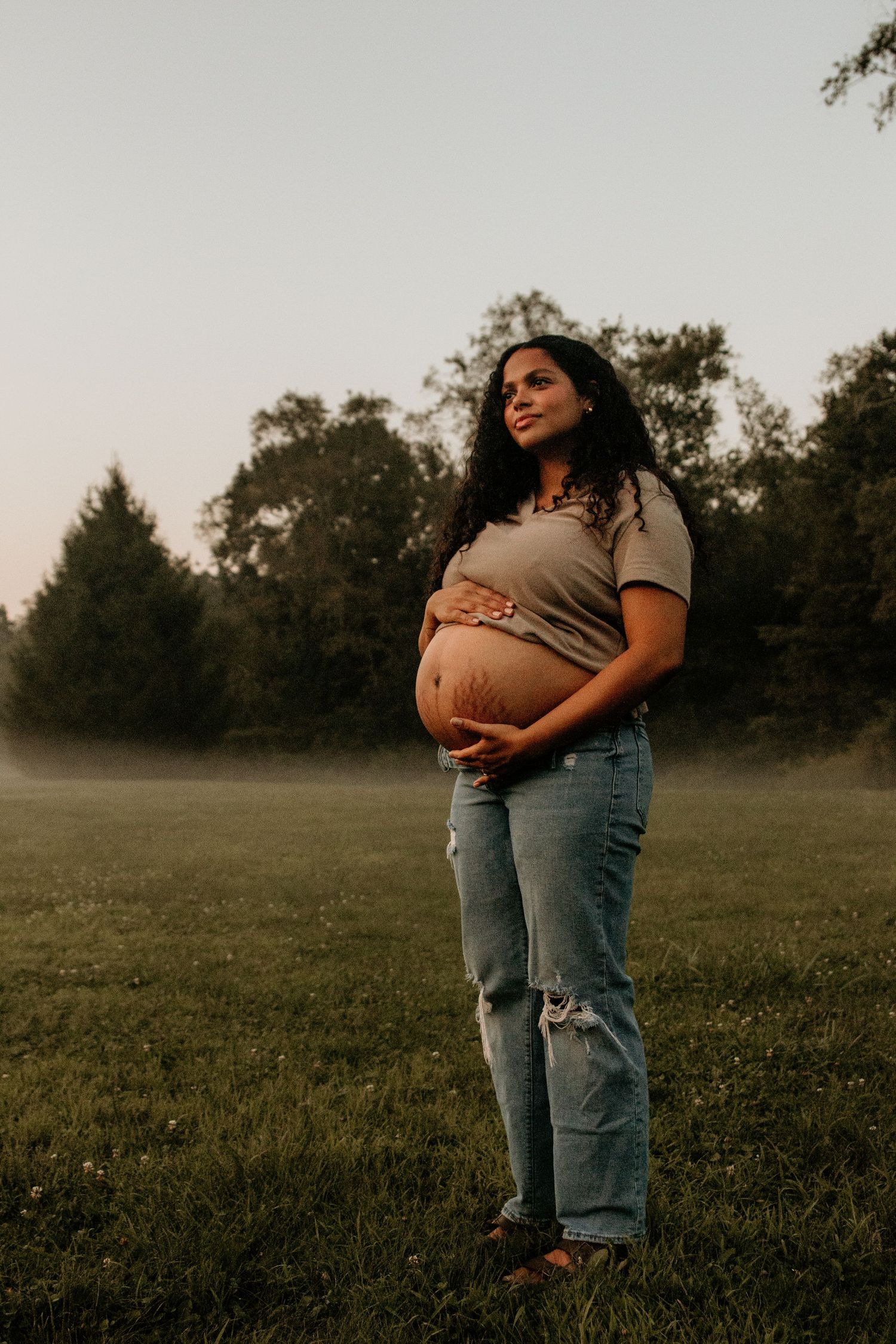 Expectant mother cradles baby bump in misty field at sunset.