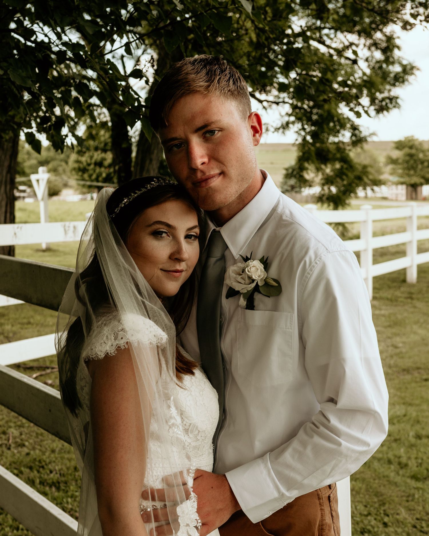 Romantic wedding portraits captured against white fence and sunlit countryside setting.