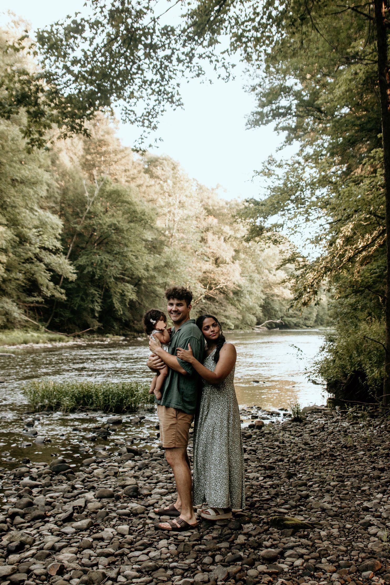 Family standing together on rocky riverbank in scenic forest setting.