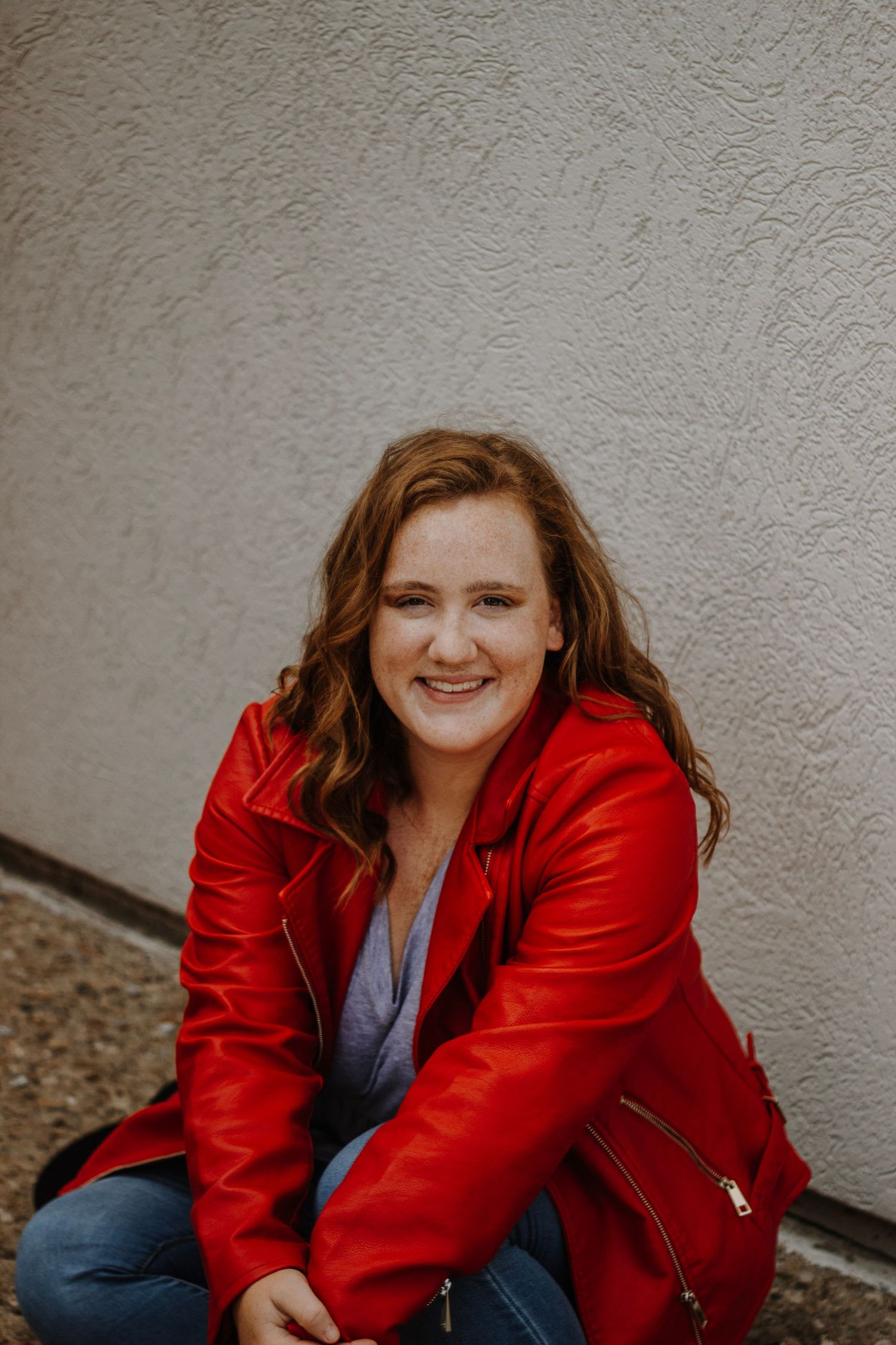 Woman in bright red leather jacket and jeans poses casually against textured wall.