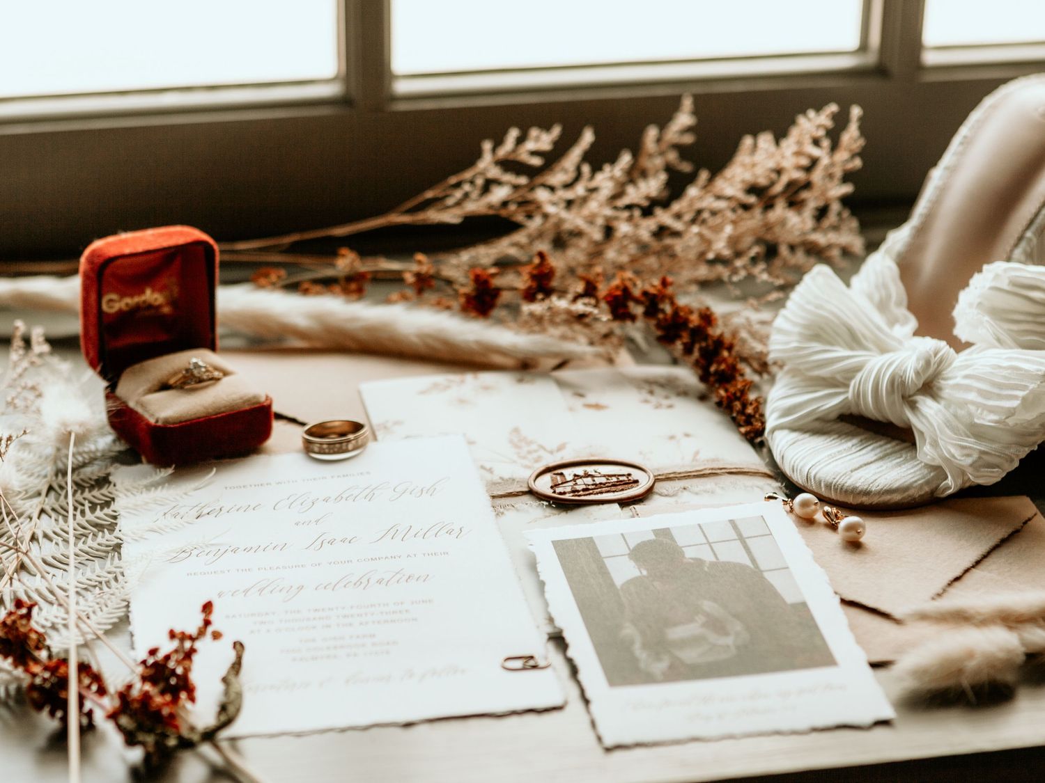 Wedding details flatlay with rings, invitation and dried flowers by window.