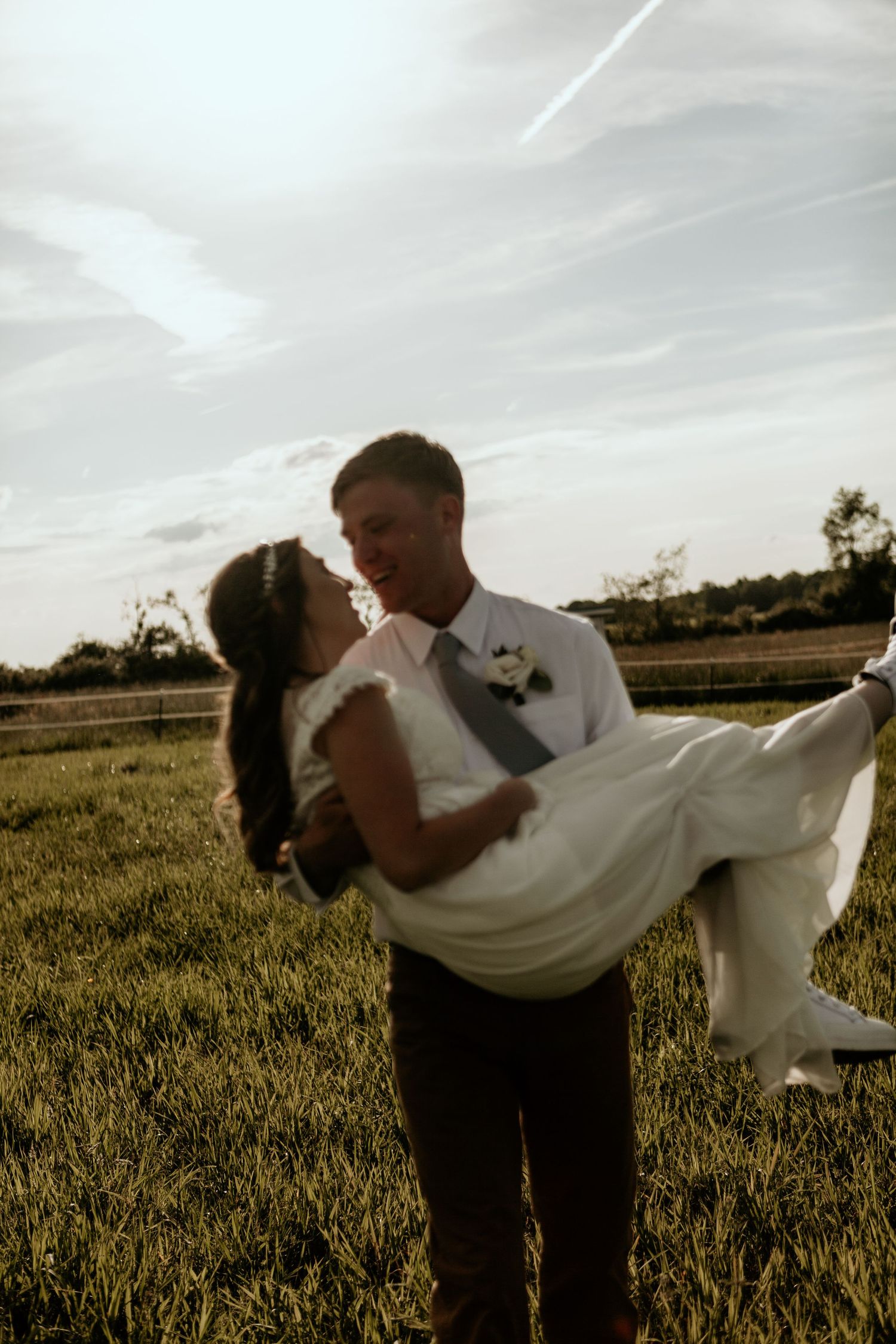 Groom carries bride across golden field during sunset wedding photoshoot.