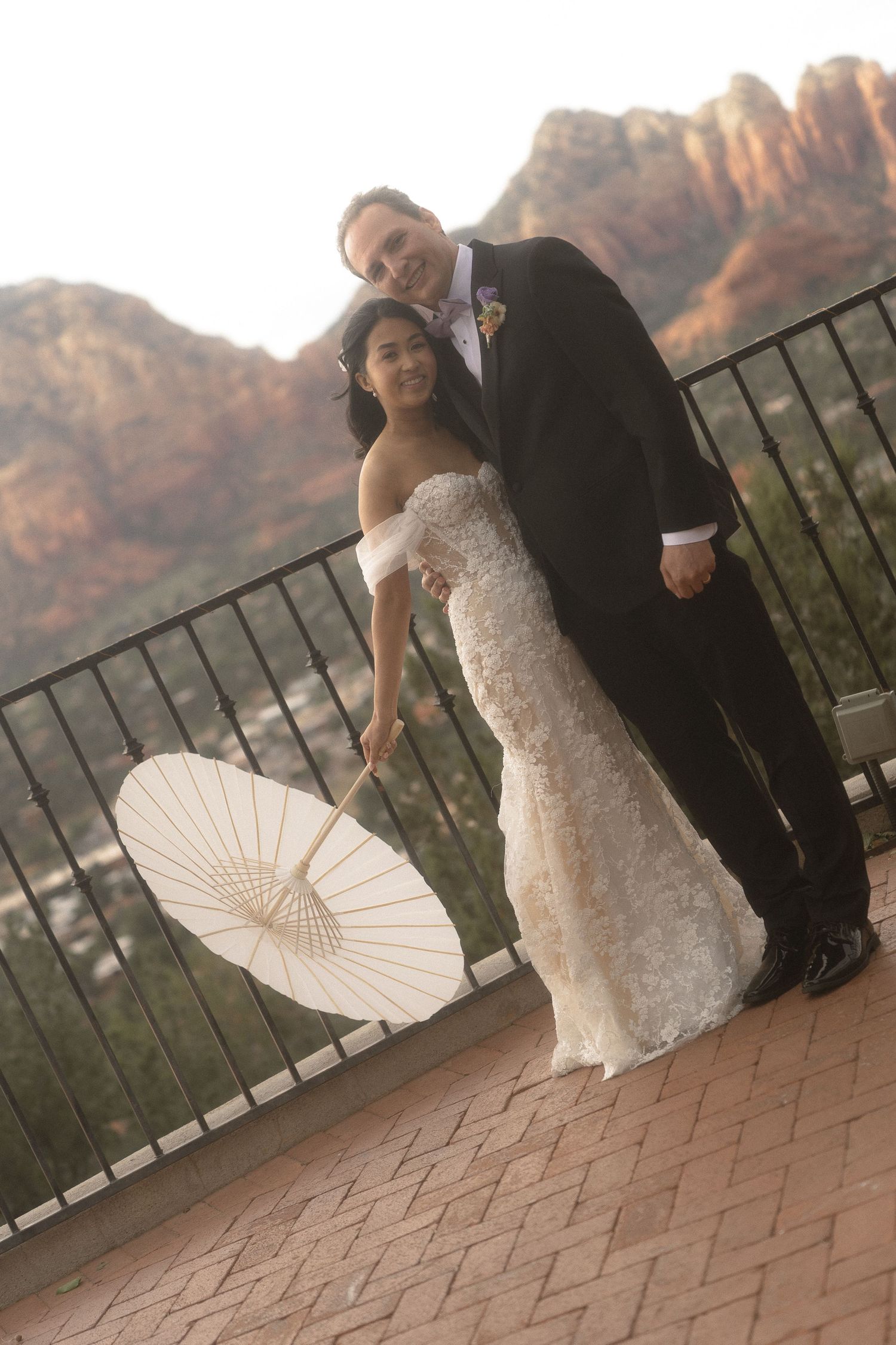 a couple stands with their backs to the railing, arizona skyline in the back, holding a pairosol