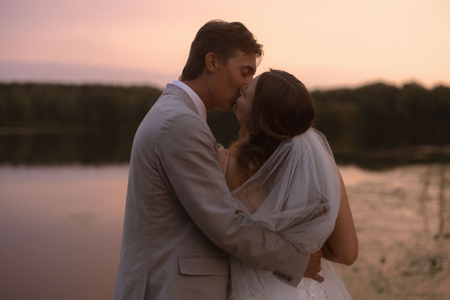a bride and groom standing by the lake at sunset.