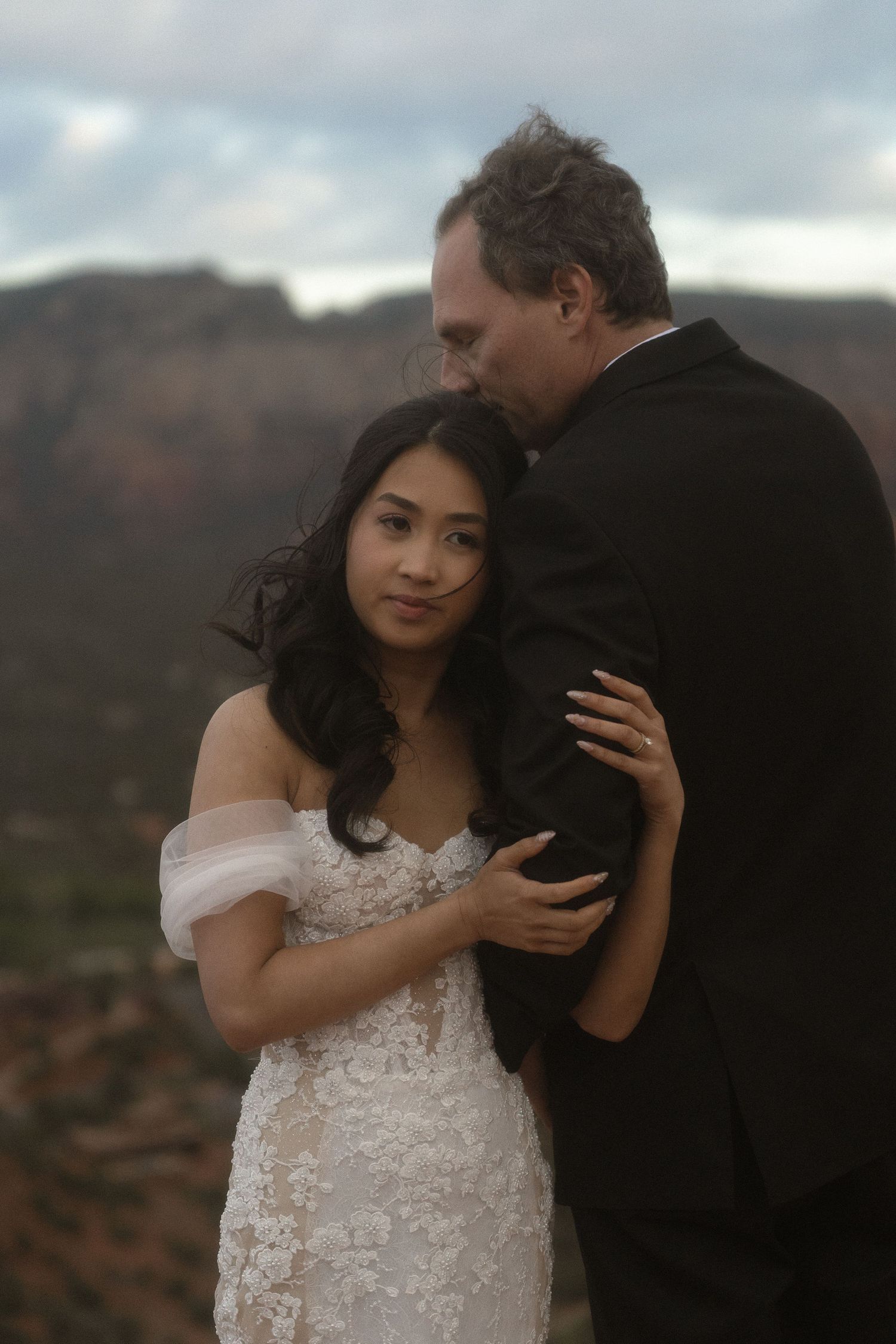 a bride hugs her husbands arm while he kisses her on the head, arizona skyline behind them