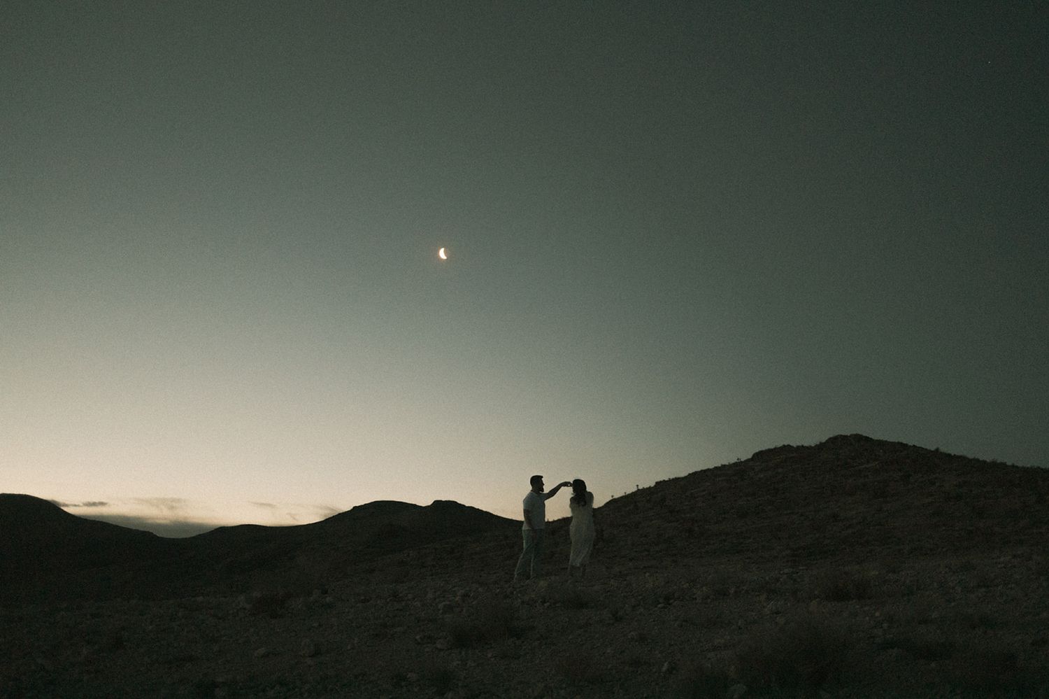 a couple dances on the nevada skyline at red rocks before sunrise, crescent moon, mountains