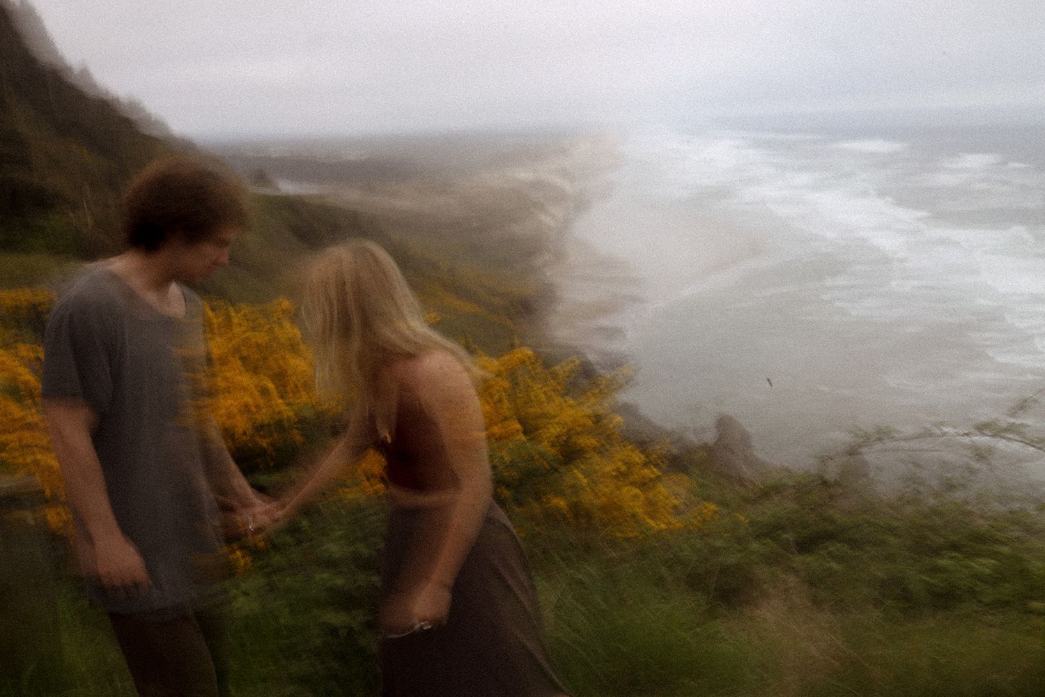 a couple stands on the oregon cliffs overlooking the pacific ocean, yellow flowers bloom over the cliffside, ethereal, motion blur