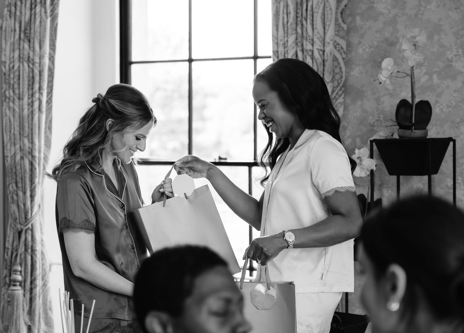 Two women exchanging gift bags indoors in a black and white photo at a elegant event.