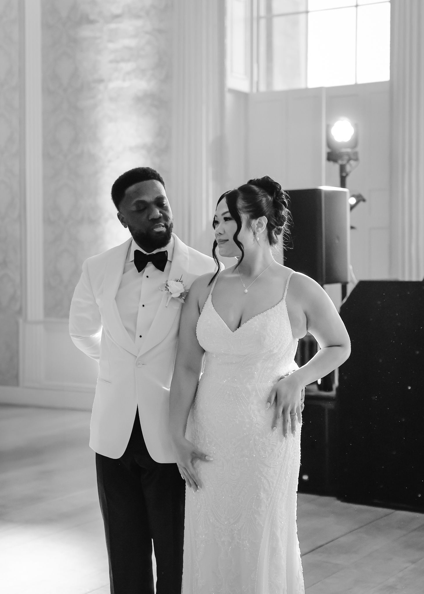 A couple in elegant wedding attire dances together in a bright, airy ballroom with white walls and natural light.