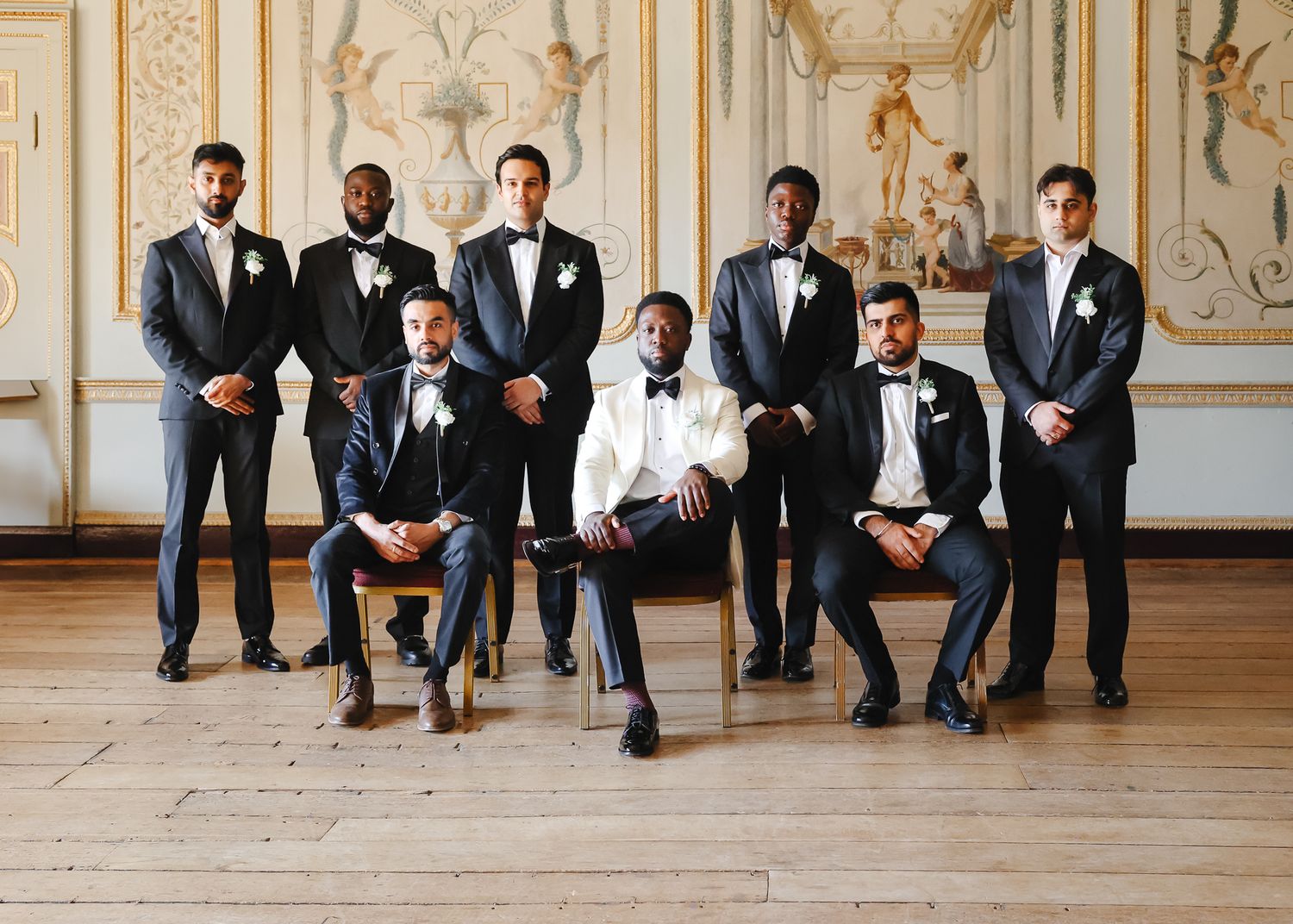 Groomsmen in formal black tuxedos and bowties pose elegantly in an ornate room with gold-detailed walls.