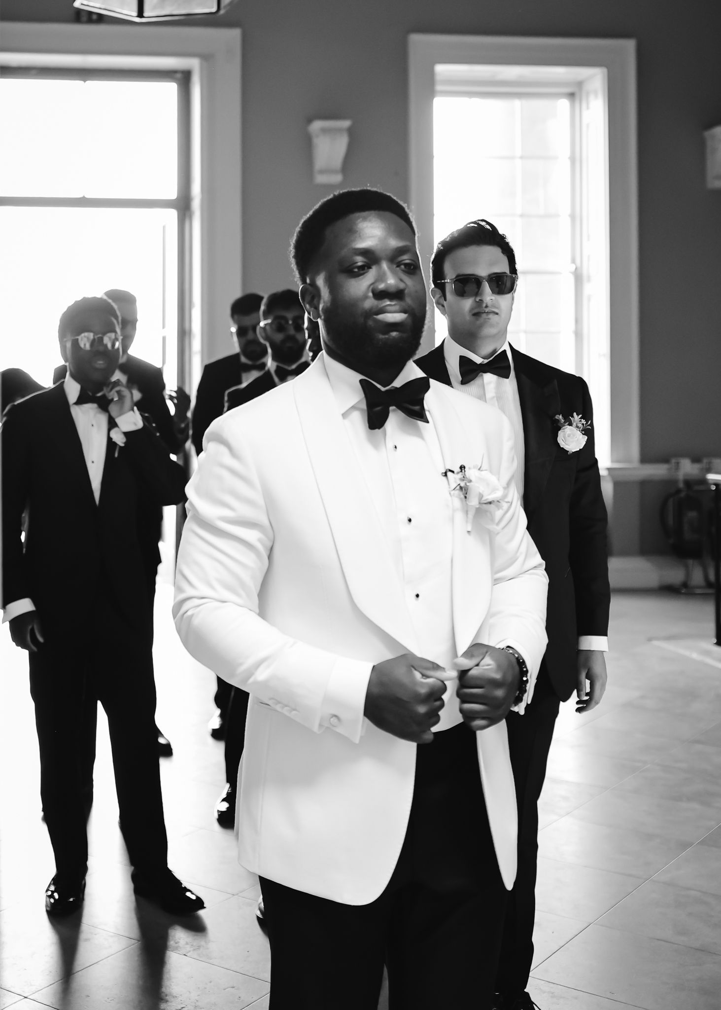 Groomsmen in black and white tuxedos walk through a bright hallway during a formal wedding event.
