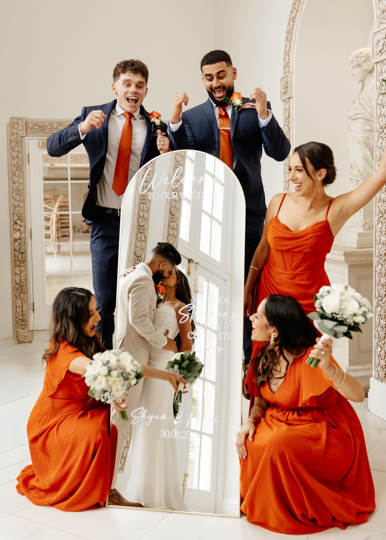Wedding party in orange bridesmaid dresses and navy suits celebrate joyfully in a bright indoor venue.
