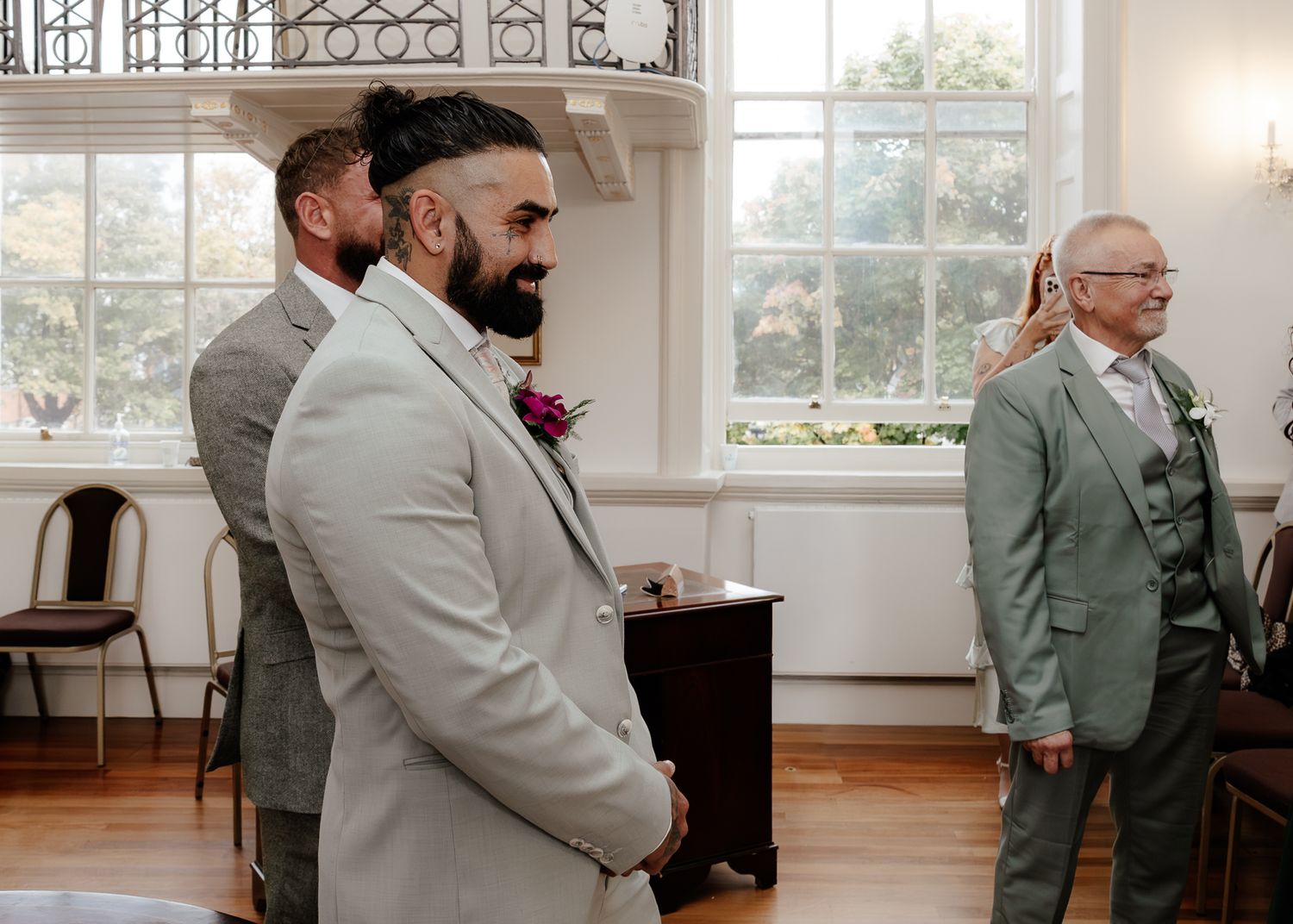 Wedding party in sage green suits stands in a bright sunlit room with white walls and large windows.