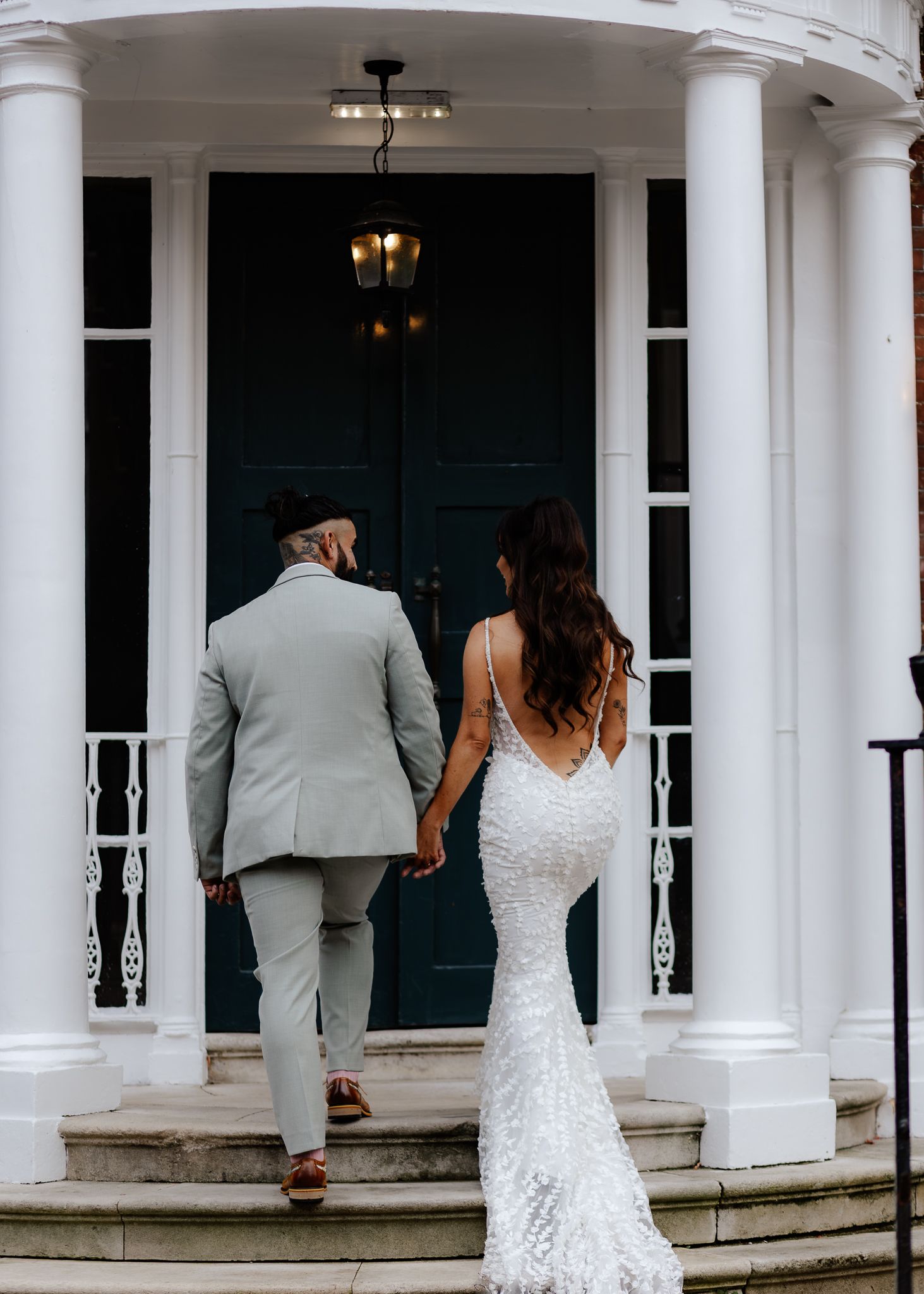 A couple in wedding attire walks along a white columned porch with elegant architectural details.