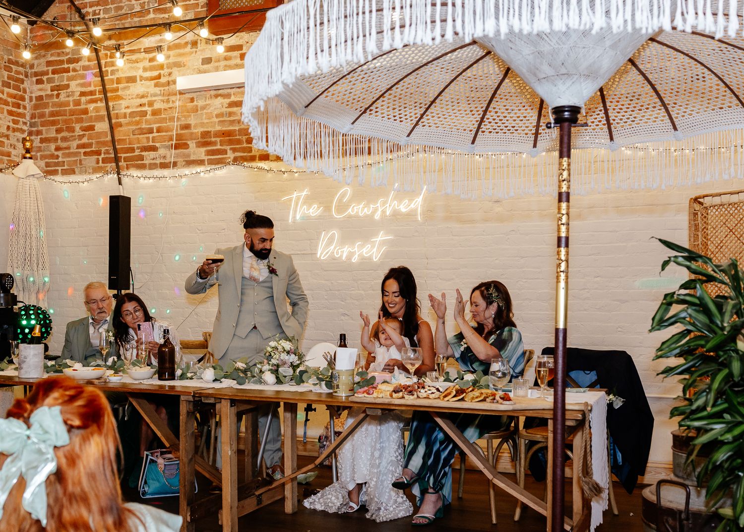 Wedding reception venue features exposed brick walls, white parasols, and elegant table settings with floral centerpieces.