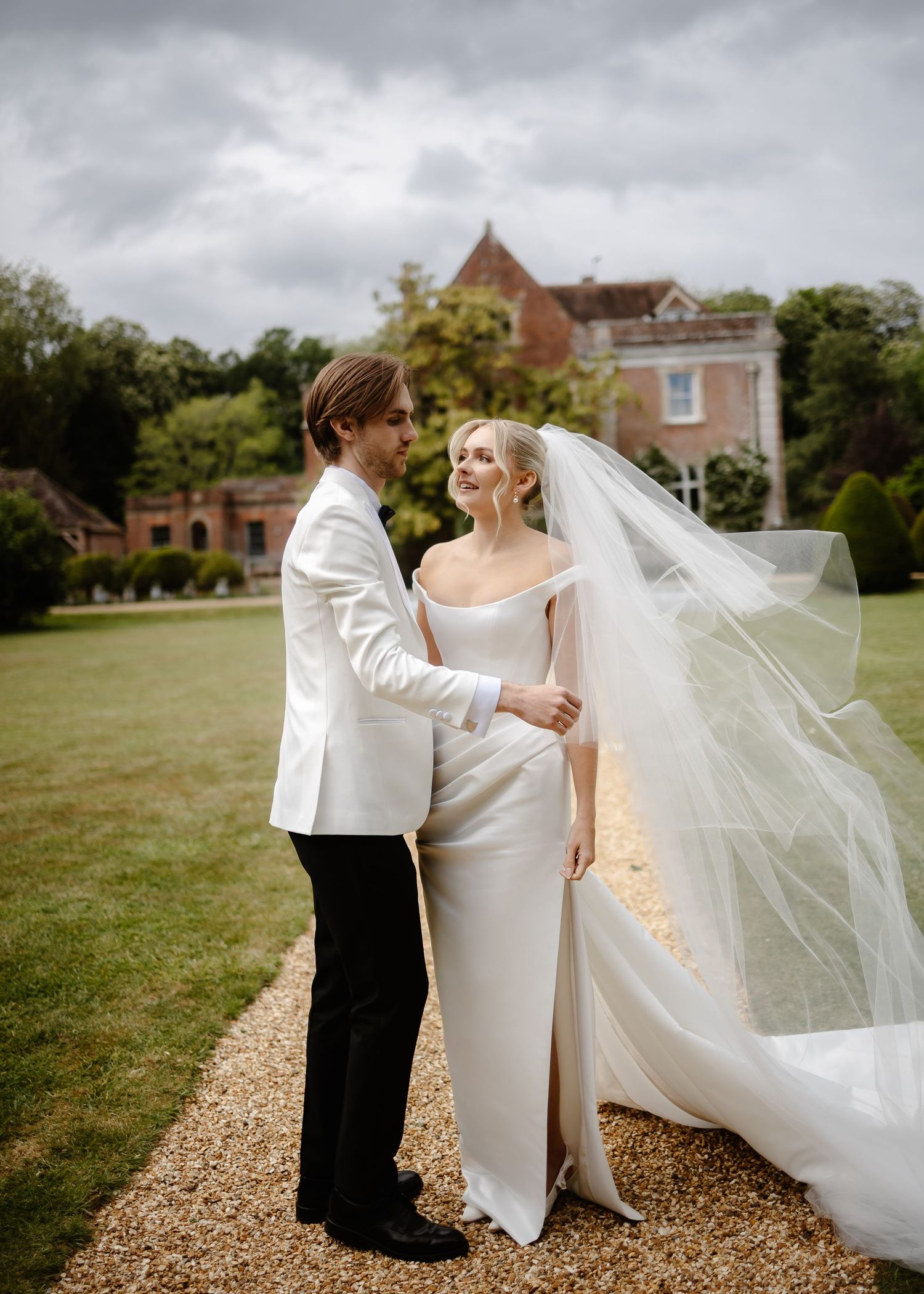 Bride and groom share a romantic moment on garden path, groom in white jacket, bride in sleek off-shoulder gown.