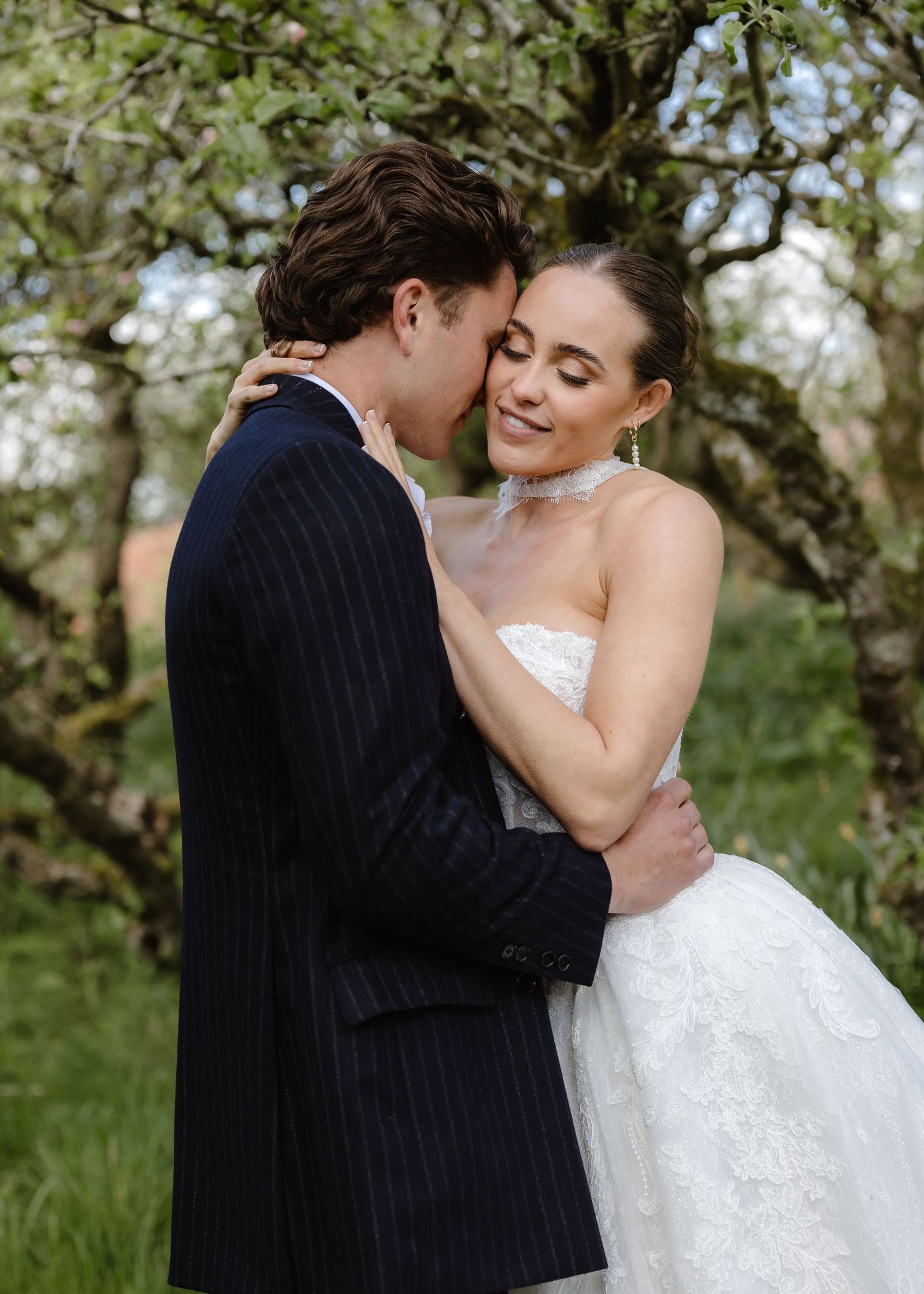 A bride and groom embrace tenderly in a lush garden, she in a strapless lace gown and he in a navy pinstripe suit.