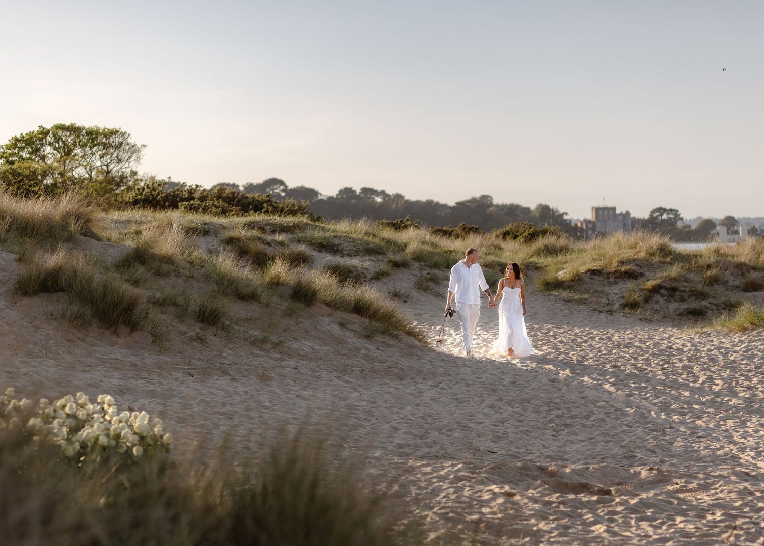 A couple in white attire walks hand-in-hand along a sandy beach path surrounded by dunes at sunset.