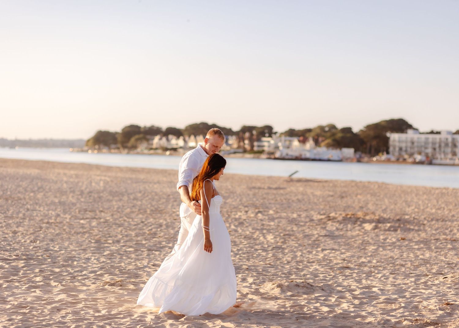 Romantic couple dancing on a sunlit beach at golden hour, woman in white maxi dress, waterfront town in background.