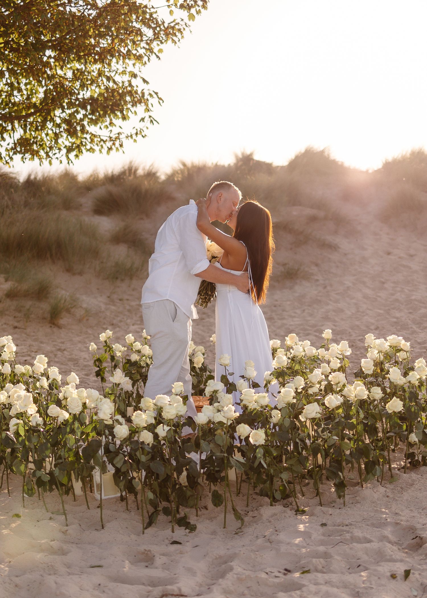 Couple kissing at sunset on sandy beach surrounded by heart-shaped arrangement of white roses during romantic proposal.