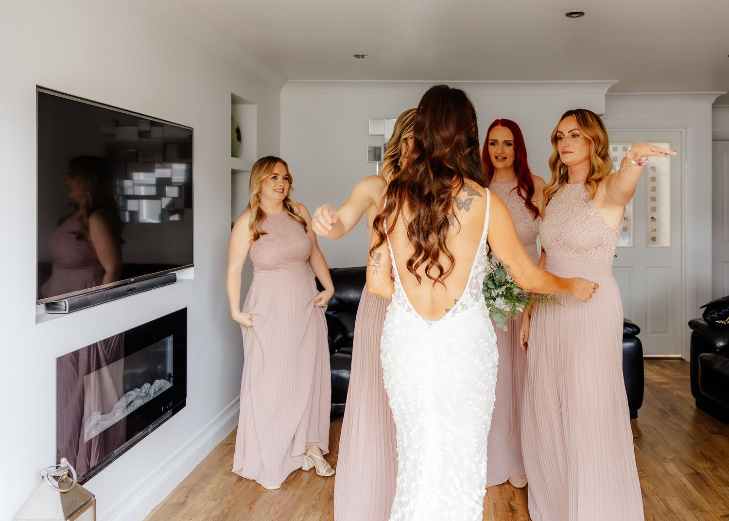 A group in matching blush pink bridesmaid dresses and a bride in white dance together in a bright modern room.