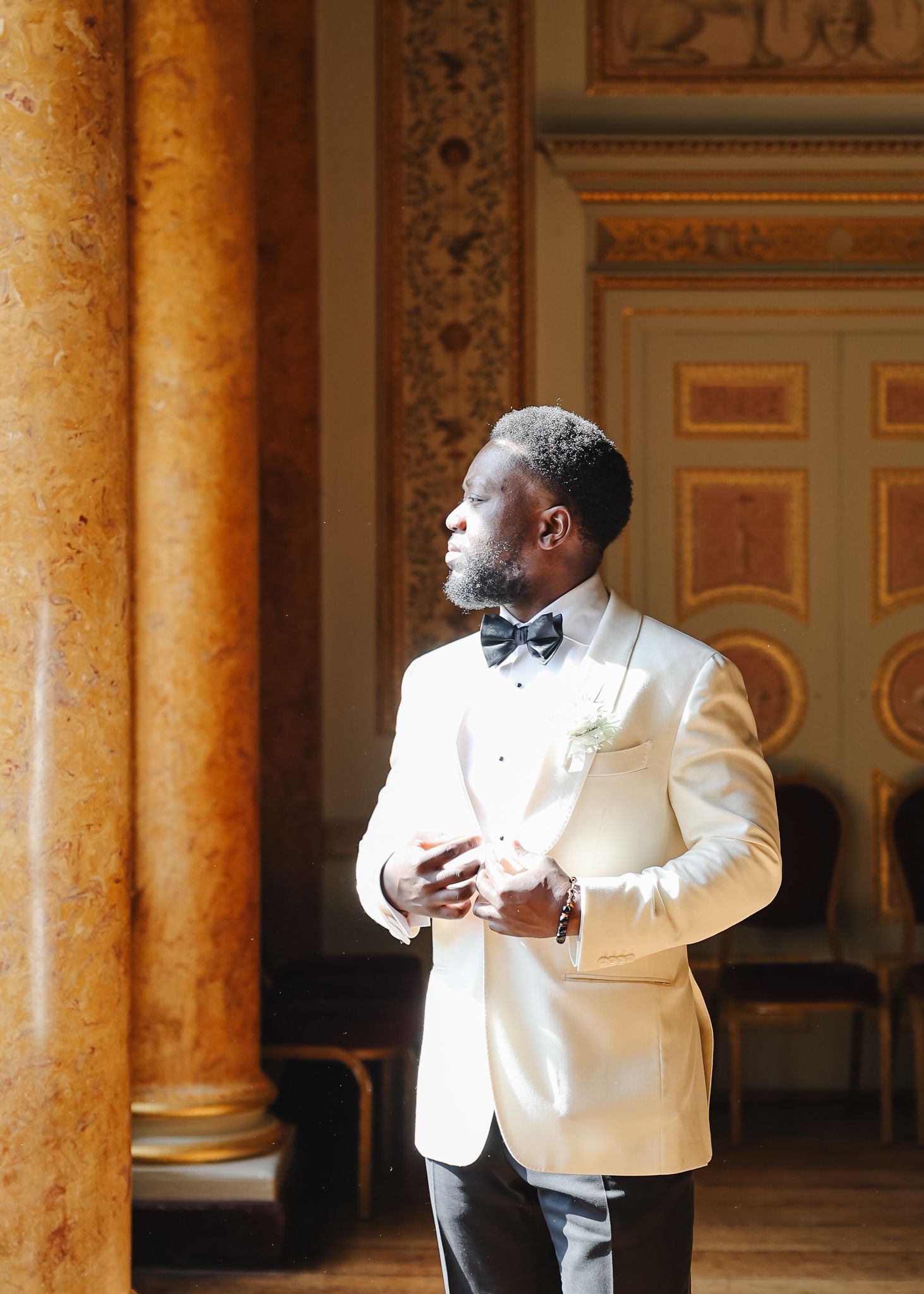 Groom in white tuxedo jacket and black bow tie poses in an ornate ballroom with marble columns.