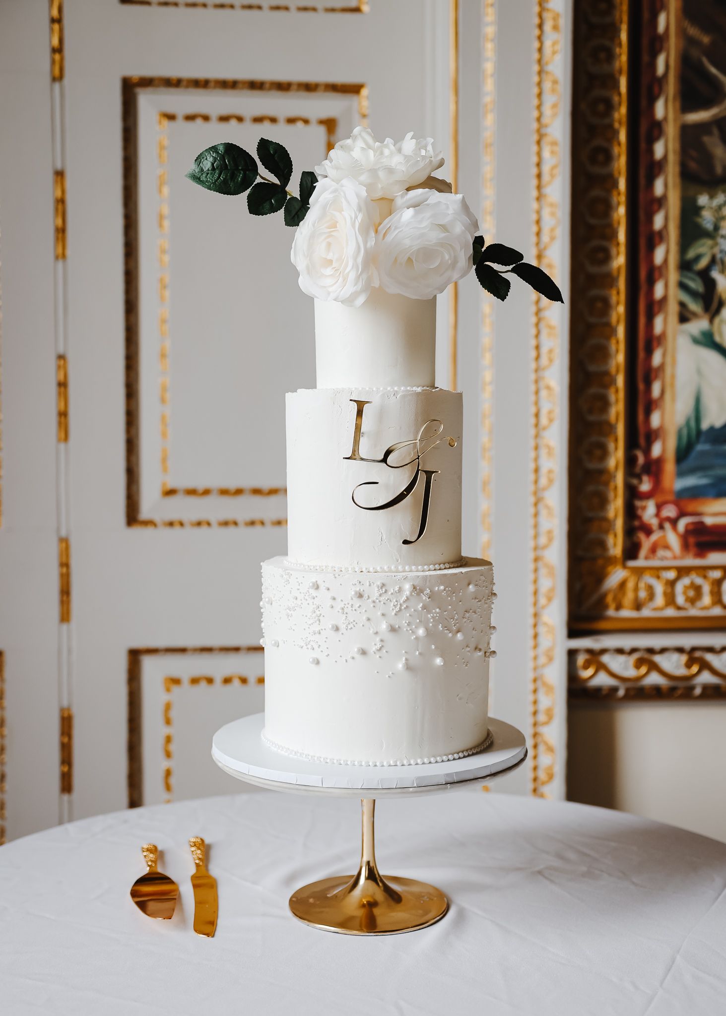 White tiered wedding cake with floral decorations displayed on a gold stand against an elegant backdrop.