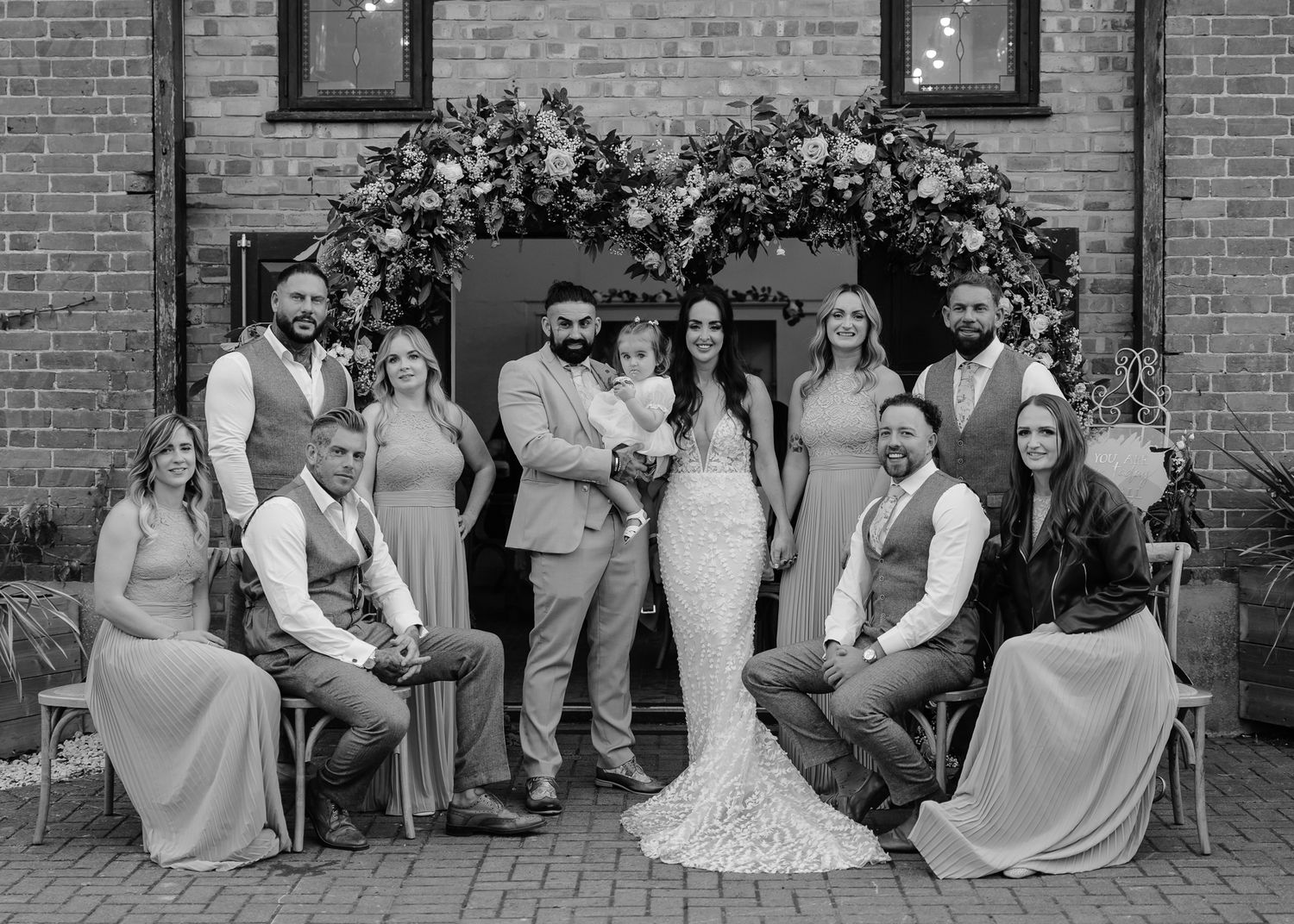 Wedding group strikes fun poses in formal attire outside a brick building.