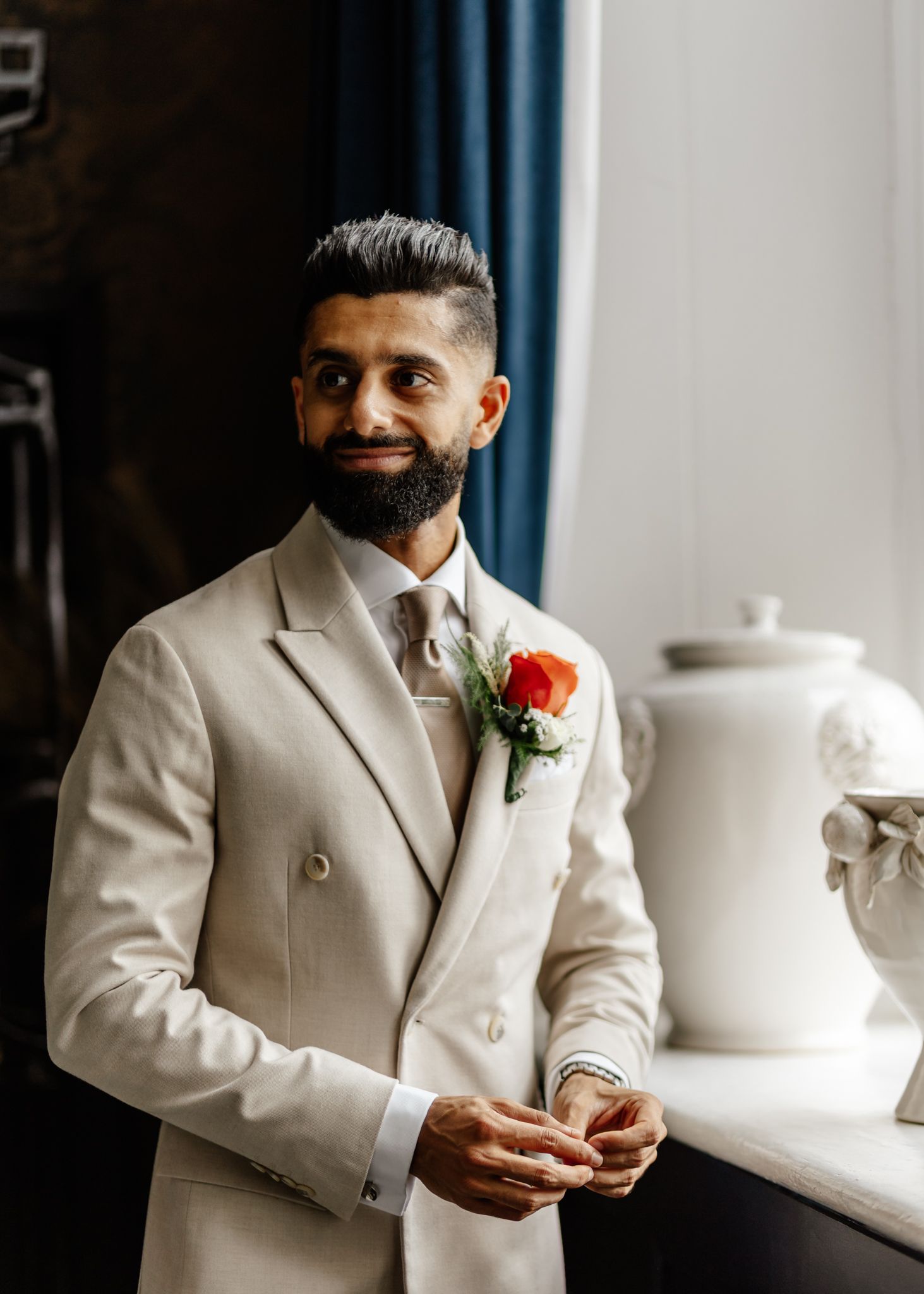 A person in a cream-colored suit with a red boutonniere holds a glass while sitting in an elegant setting.