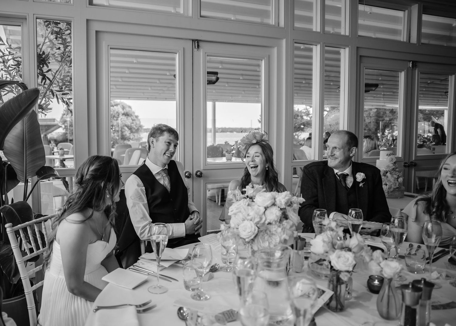 Black and white wedding reception photo of guests laughing at a beautifully decorated table with floral centerpieces.