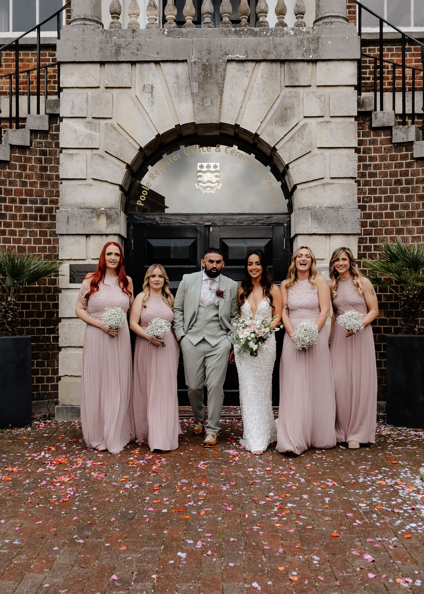 Wedding party poses under stone archway with bride in white and bridesmaids in blush pink dresses with flower petals scattered.