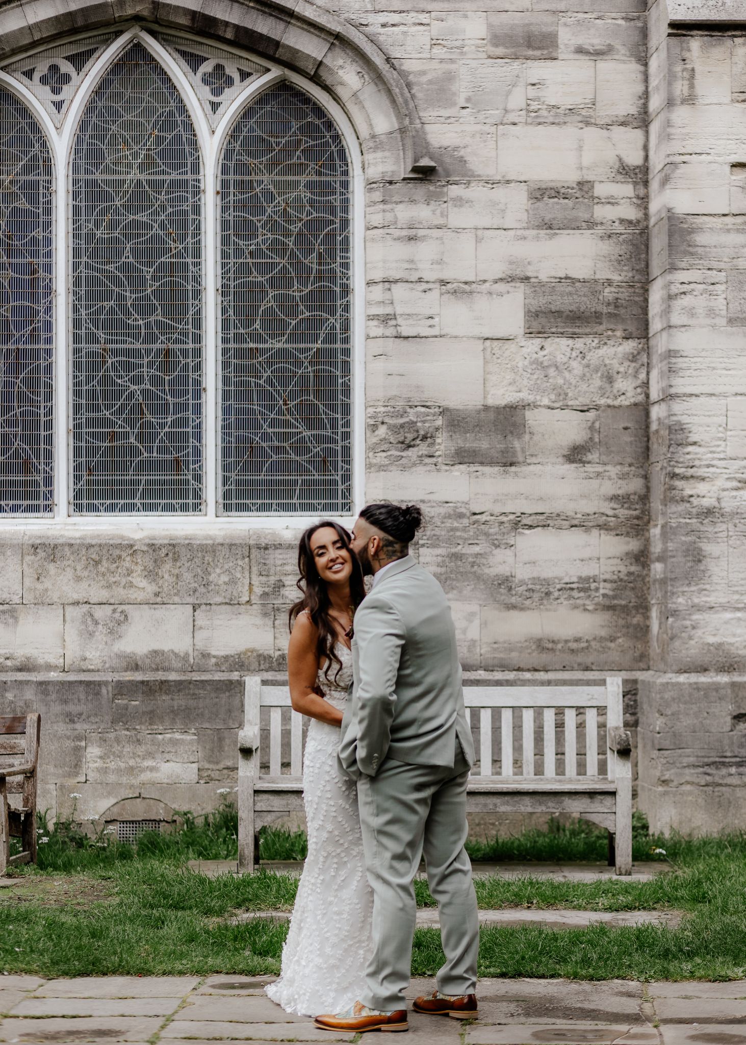 A couple shares a joyful moment in their wedding attire outside a historic stone church with gothic windows.