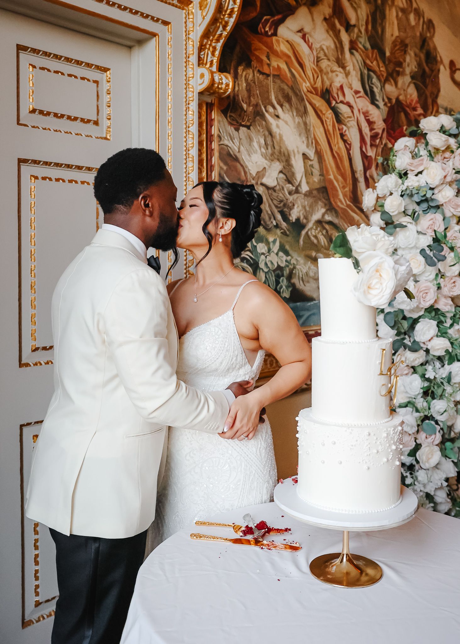 Elegant white tiered wedding cake displayed in front of ornate wall with floral decorations.