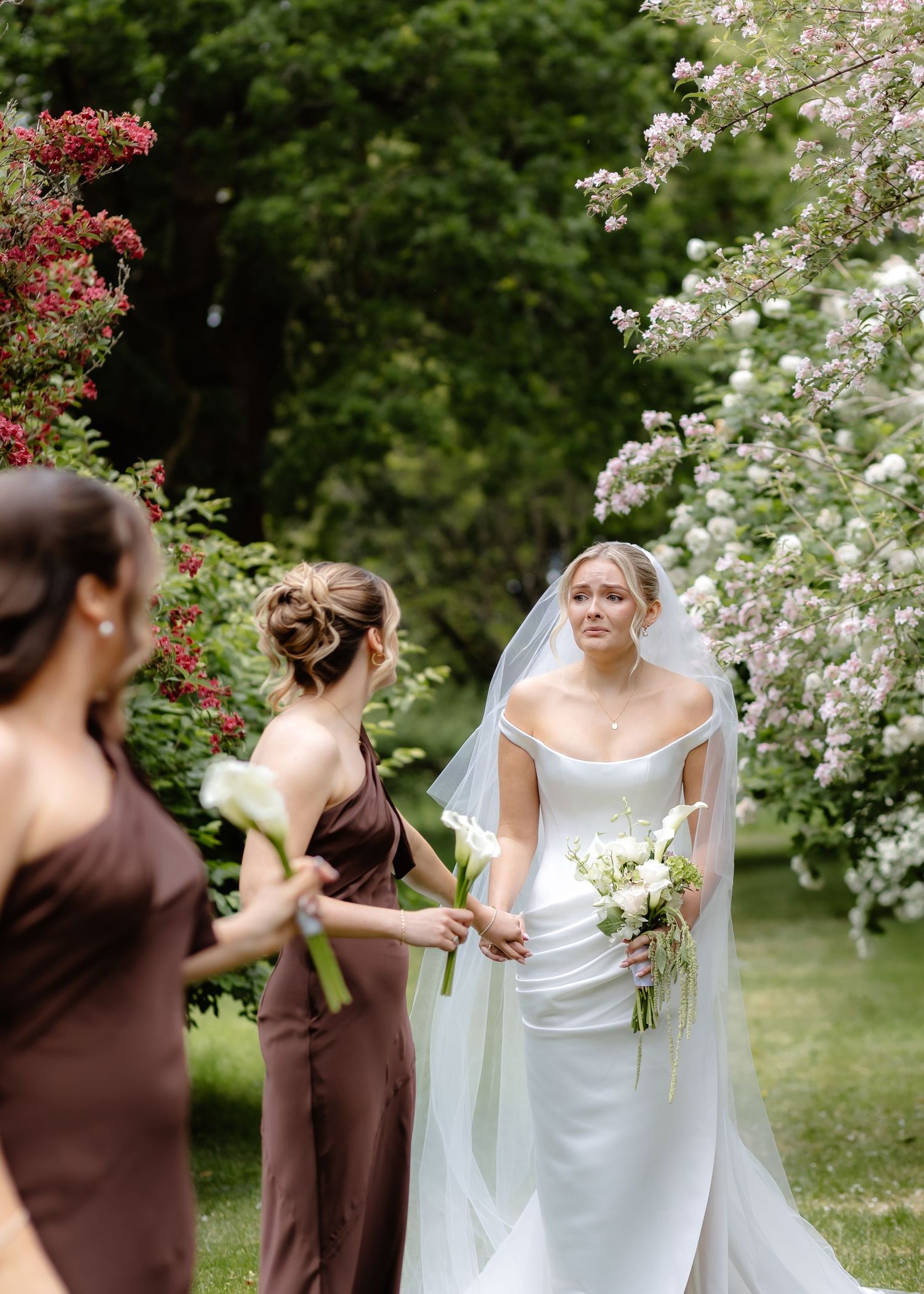 Bride in white gown and veil holds bouquet with bridesmaids in brown dresses in a lush garden setting.