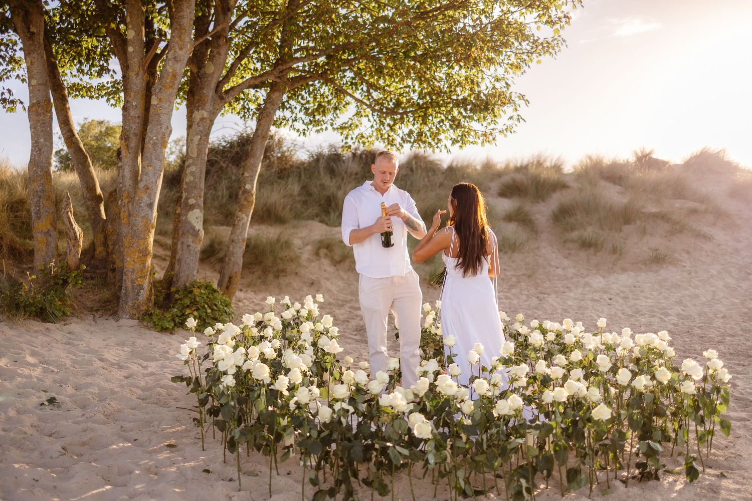 Couple in white outfits sharing a romantic beach proposal moment surrounded by white roses at golden hour.