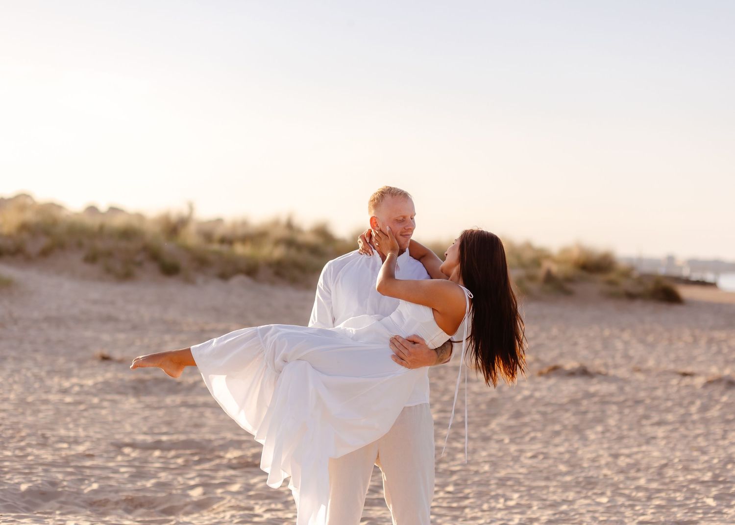 A man in white lifts a woman in a white dress on a sandy beach at sunset, sharing a romantic moment.