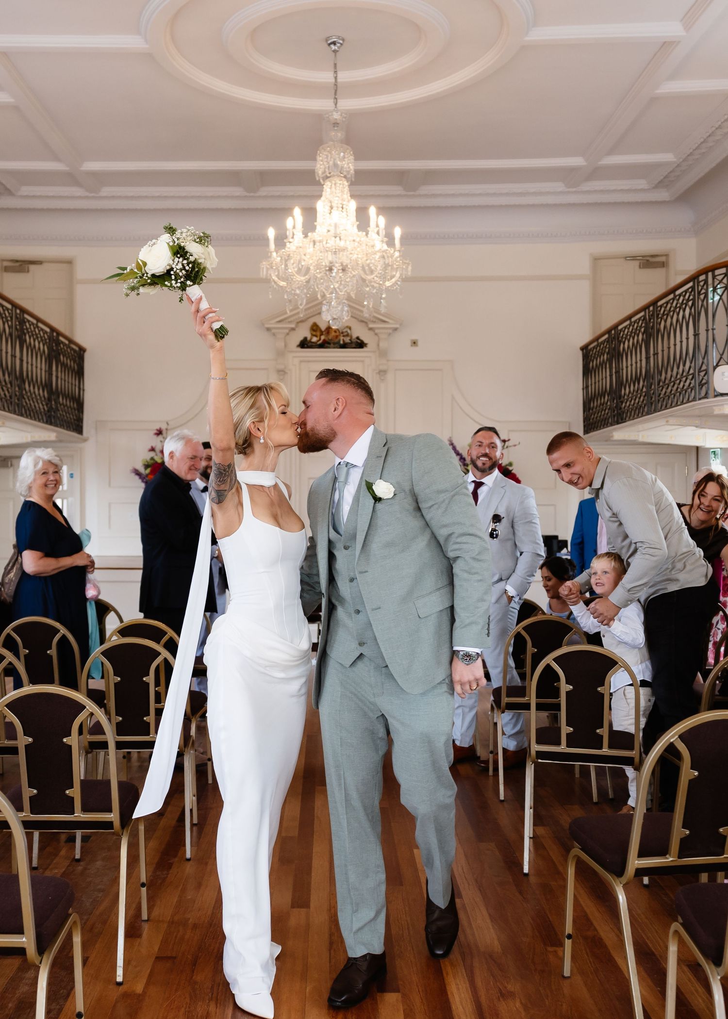 Newlyweds share a kiss while bride raises bouquet in elegant chandelier ceremony hall with guests celebrating.