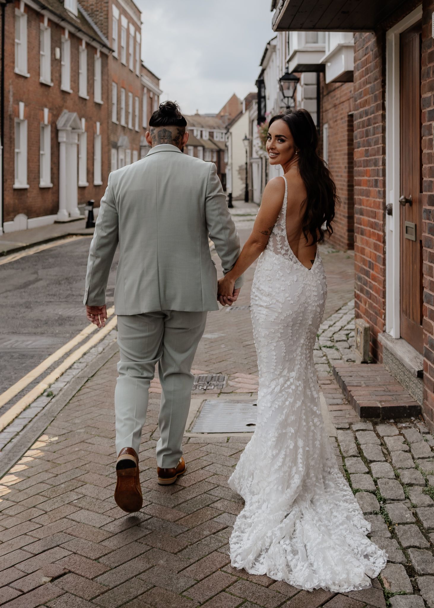 A couple walks together on a cobblestone street, with the bride wearing a lace wedding dress and groom in sage green suit.